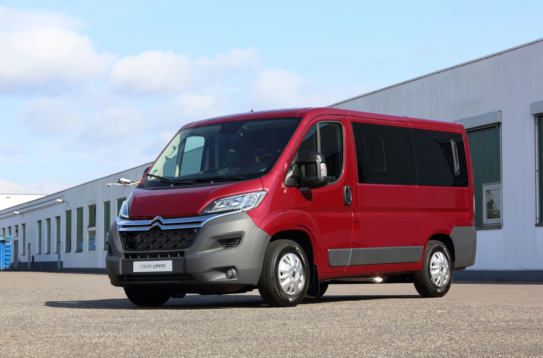 Roter Citroën Lieferwagen steht auf einem asphaltierten Platz vor einem großen, weißen Industriegebäude unter blauem Himmel.