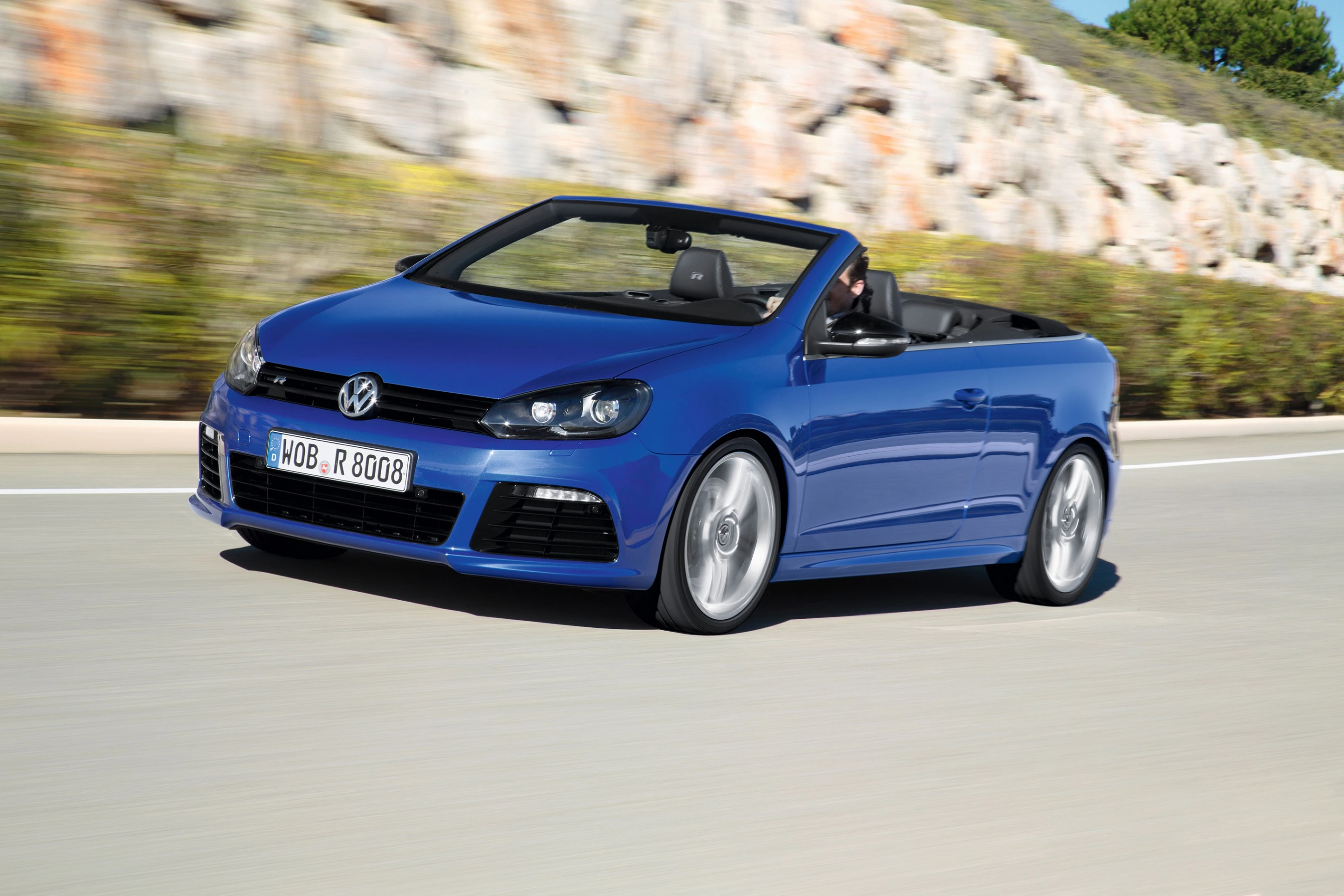 Blaues VW-Cabriolet fährt auf einer Straße vor einer Steinmauer. Fahrer sitzt im Auto, Himmel klar.