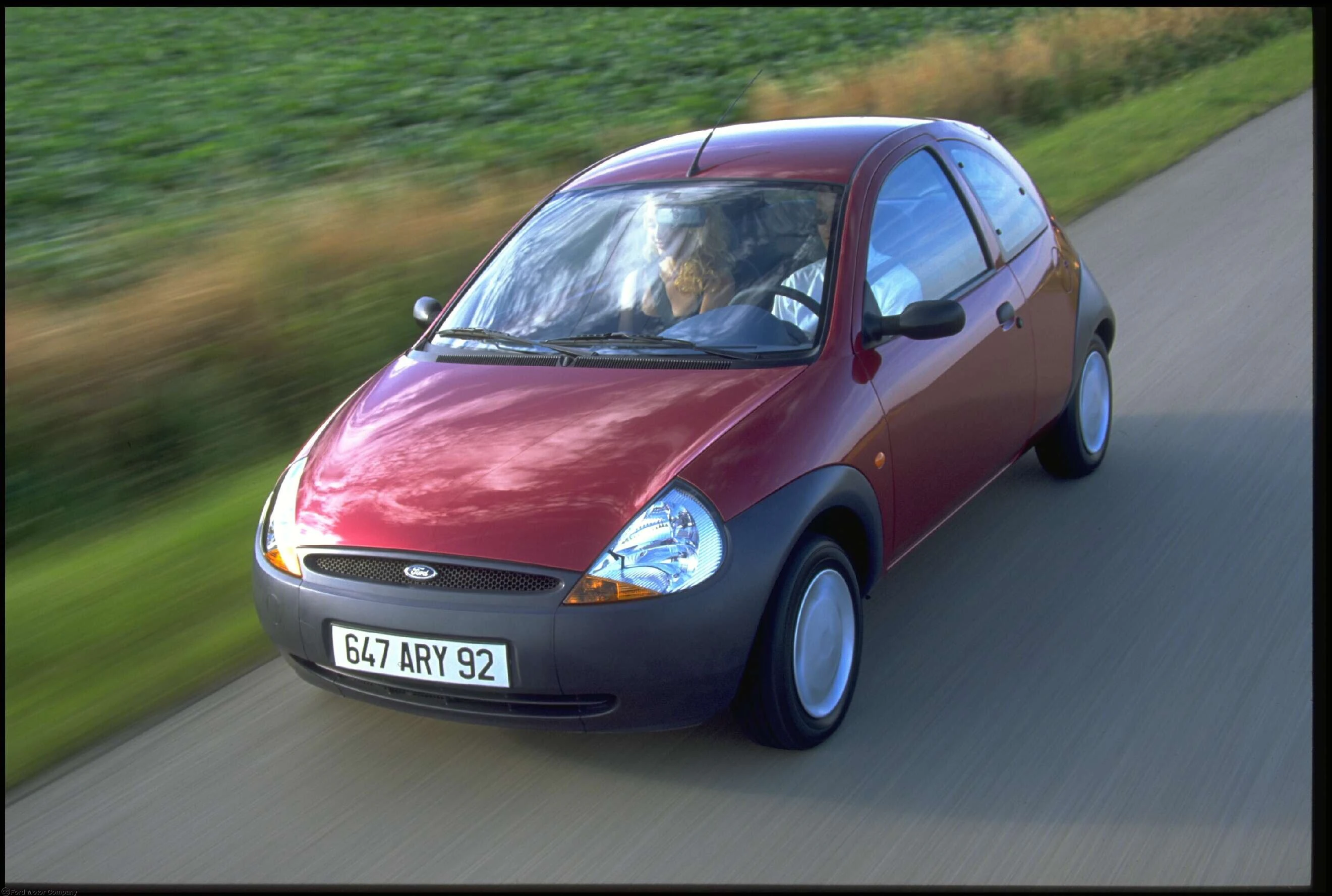 Roter Ford Ka fährt auf einer Landstraße, umgeben von grüner Landschaft. Ansicht von schräg oben, Fahrer sichtbar.