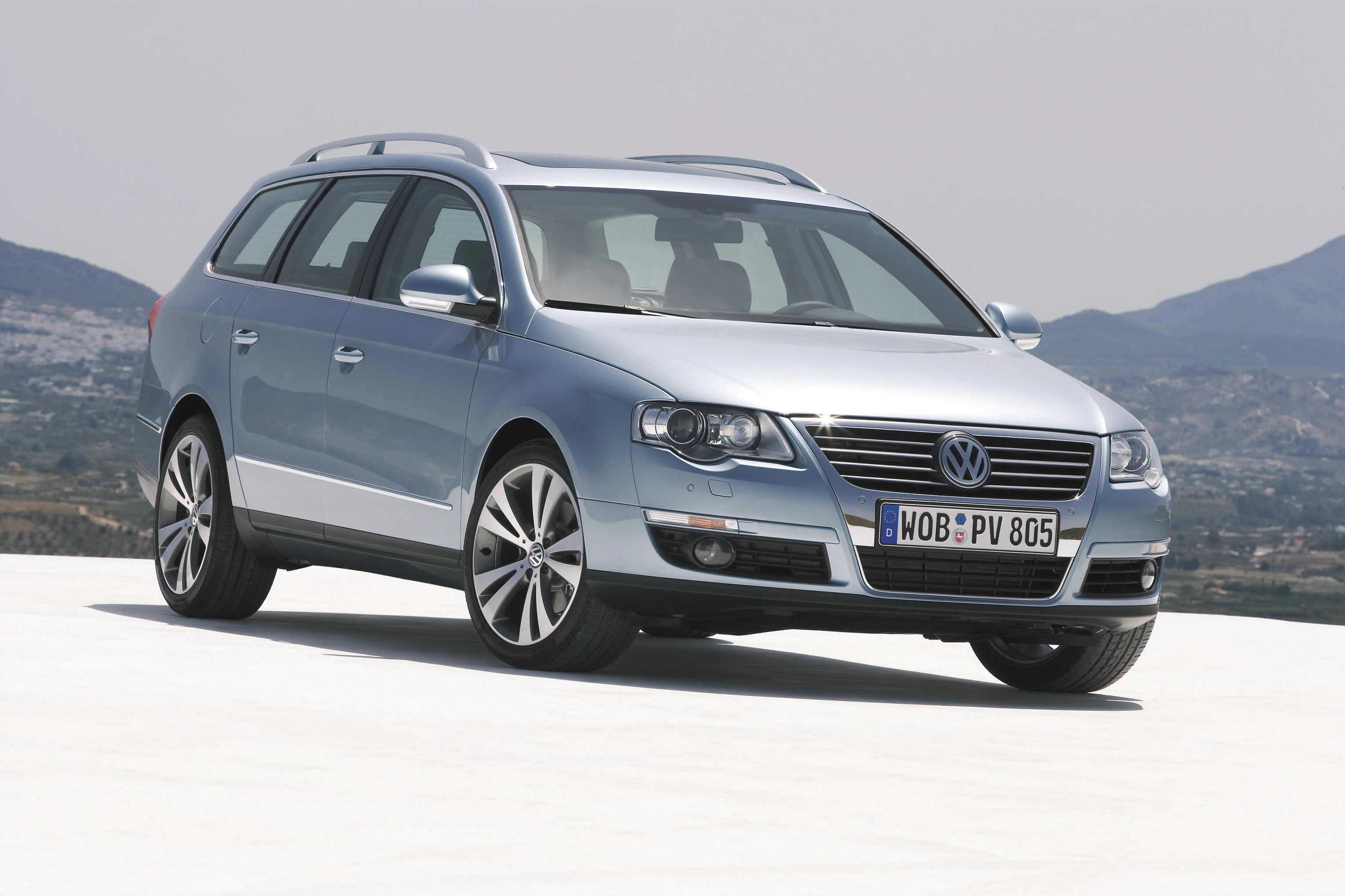 Silberner Volkswagen Passat Kombi steht auf einer weißen Fläche vor einer hügeligen Landschaft bei klarem Himmel.