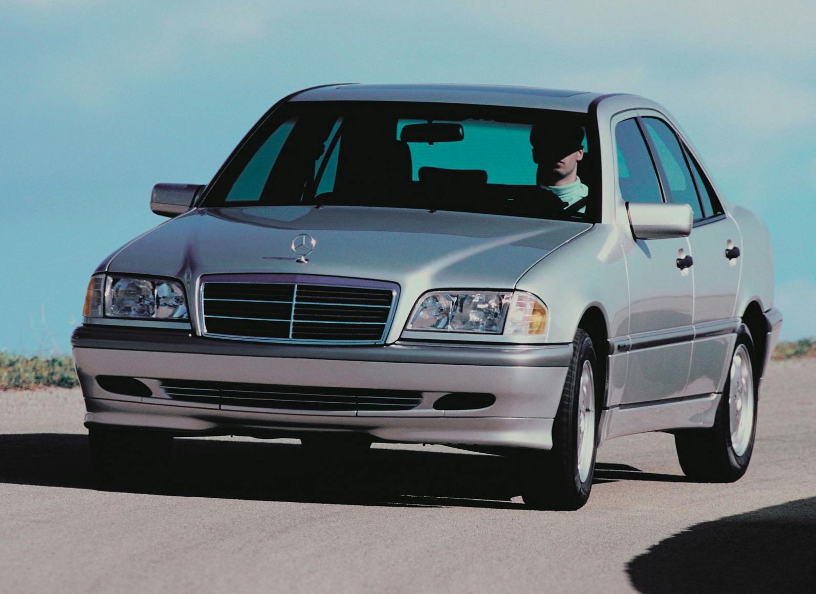 Silberner Mercedes-Benz Limousine fährt auf einer Straße, Fahrer im Innenraum sichtbar, bei sonnigem Wetter.
