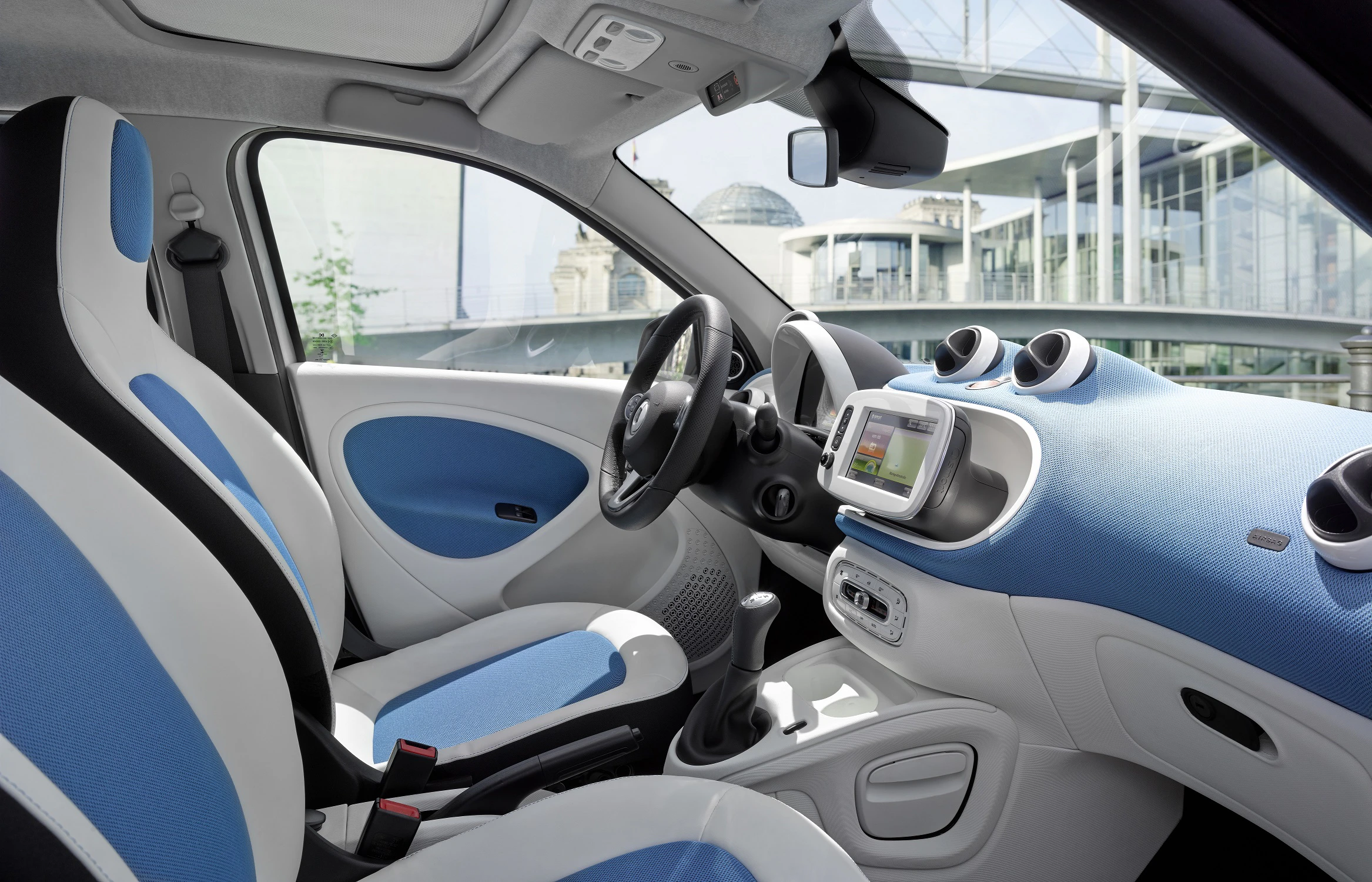Innenraum eines modernen Autos mit blau-weißen Sitzen, digitalem Armaturenbrett und Blick auf ein urbanes Gebäude durch das Fenster.