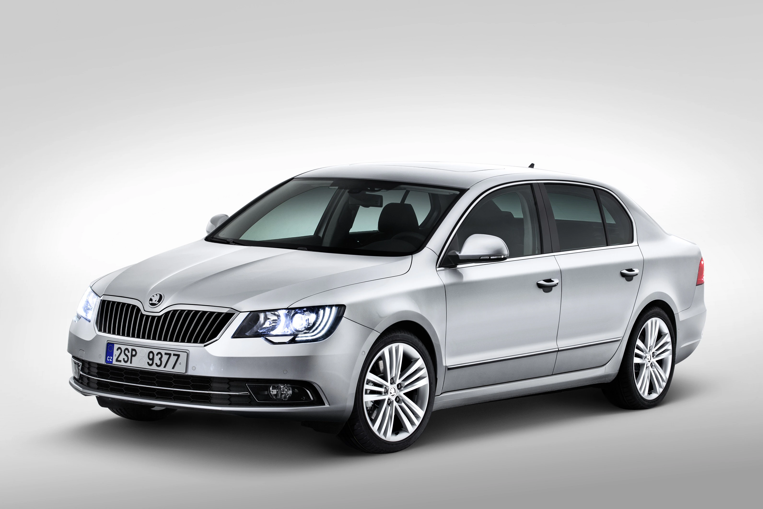 Silberner Skoda Superb Limousine vor grauem Hintergrund, Frontansicht mit eingeschalteten Scheinwerfern und tschechischem Kennzeichen.