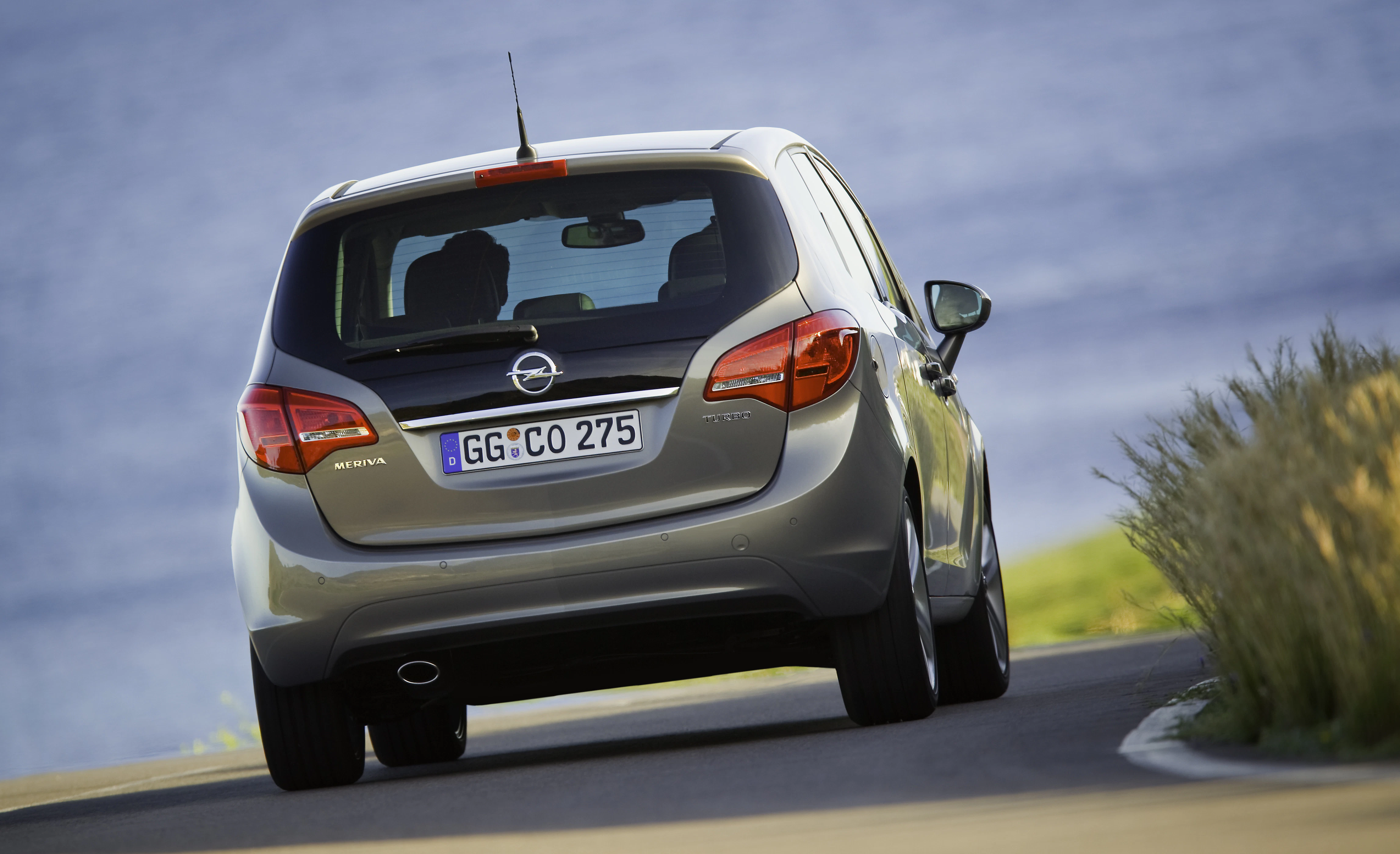 Silberner Opel Meriva auf einer kurvigen Straße am See entlang, Autoscheinwerfer aus, Wiesen im Hintergrund, Himmel blau.