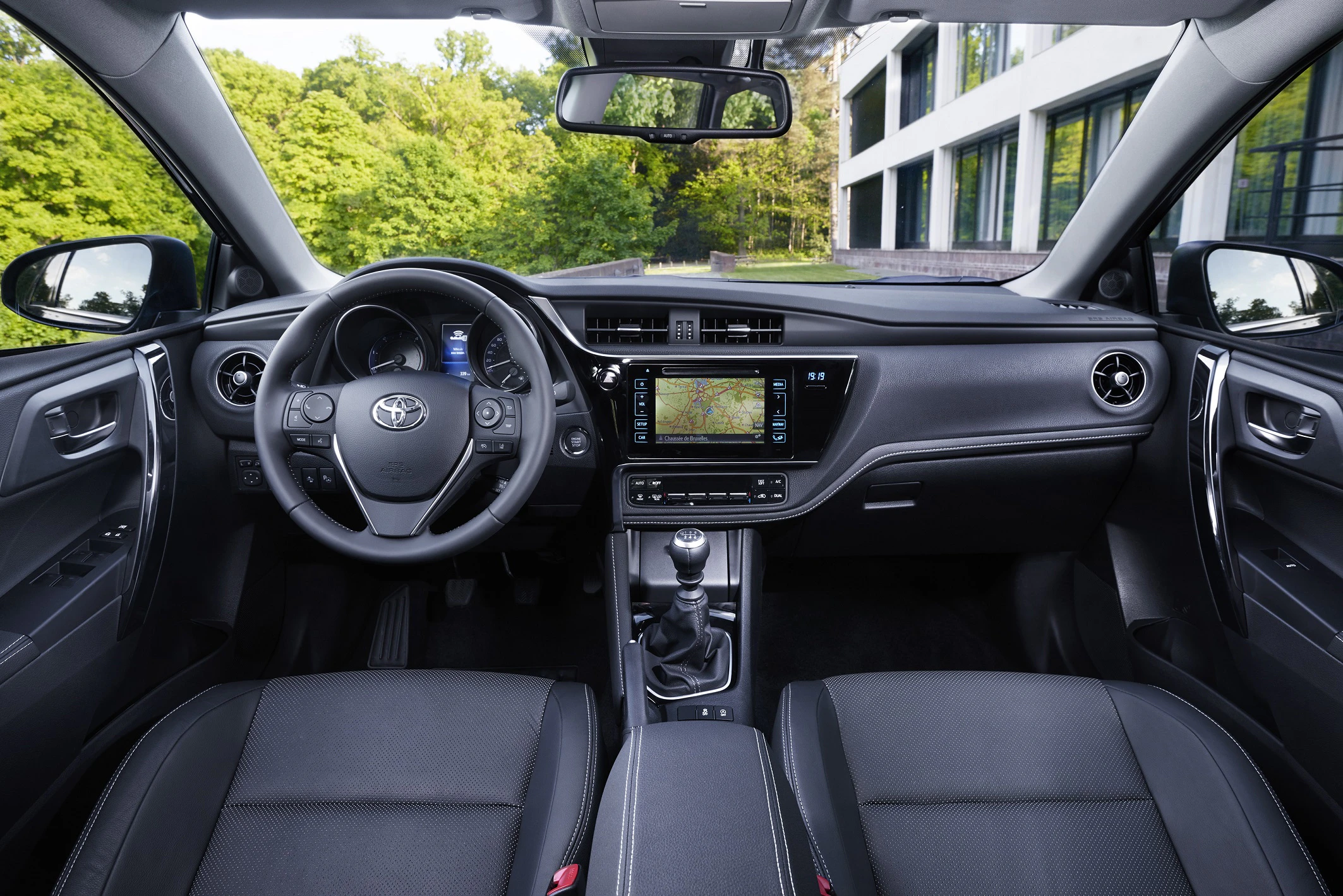 Innenraum eines Autos mit modernem Armaturenbrett, Lenkrad, Touchscreen-Anzeige und manuellem Schalthebel. Fenster mit grüner Sicht nach draußen.