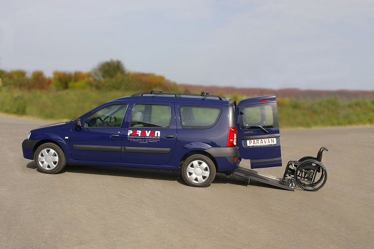 Blaues Auto mit offenem Heckklappe und Rollstuhlrampe, die zu einem Rollstuhl führt. Fahrzeug steht auf einer asphaltierten Fläche.