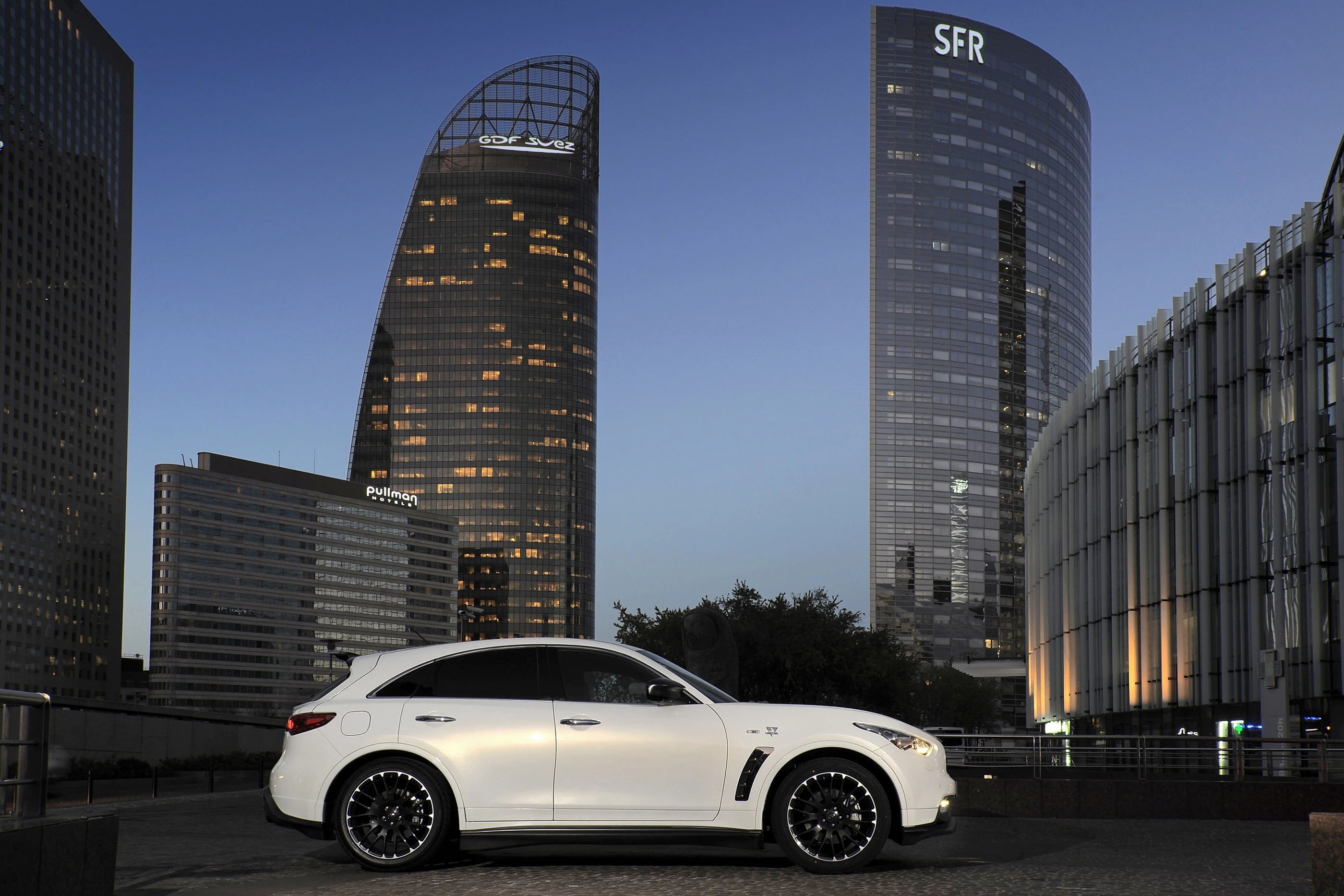 Weißes Auto vor modernen Wolkenkratzern in der Dämmerung. Im Hintergrund beleuchtete Gebäude mit Glasfassaden und das SFR-Logo.
