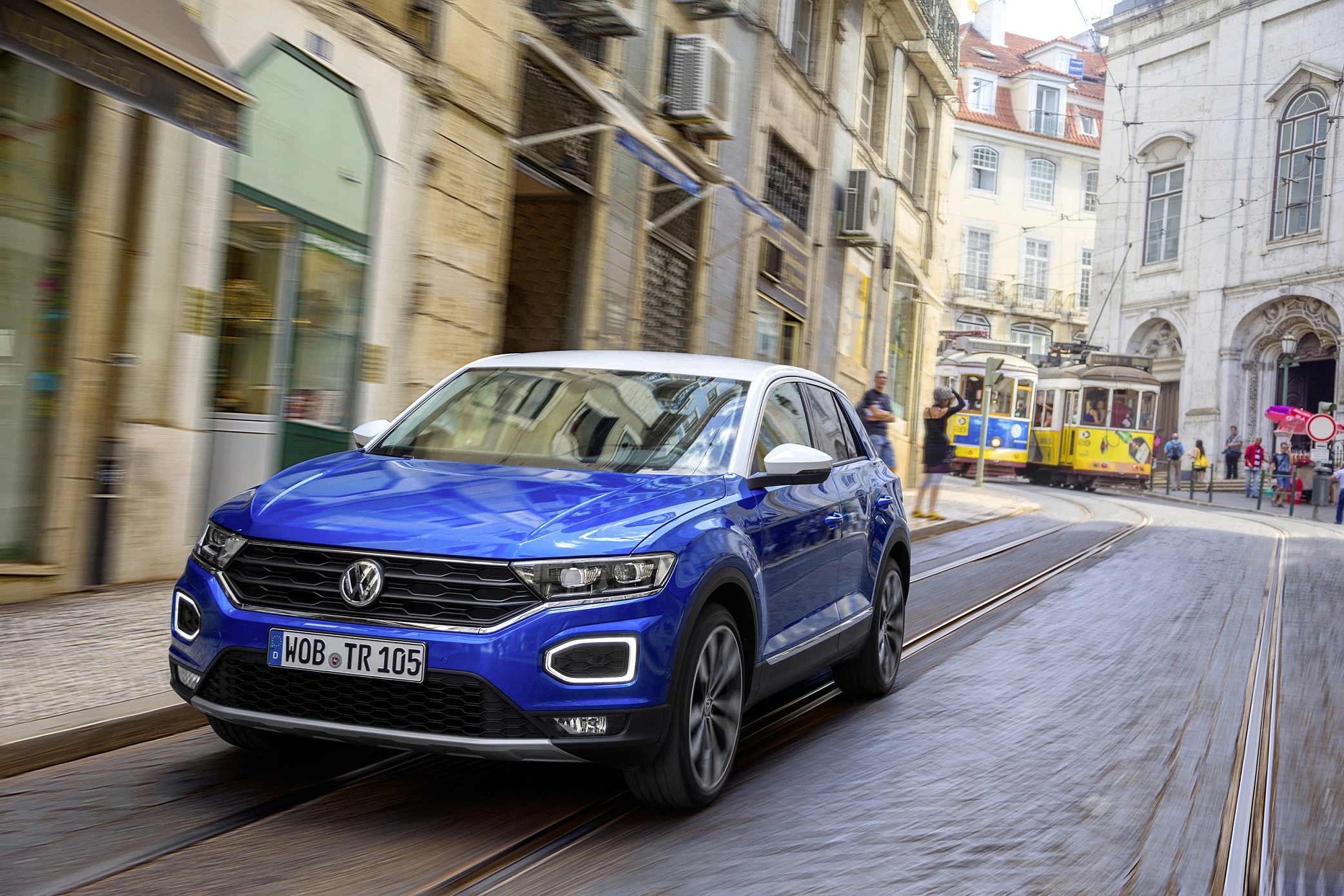 Blauer Volkswagen T-Roc fährt durch eine historische Straße mit Straßenbahn, alten Gebäuden und Passanten im Hintergrund.