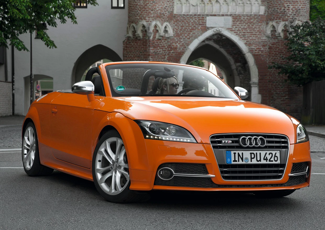 Oranger Audi TT Roadster parkt vor einem historischen Backsteingebäude. Das Verdeck ist offen, das Auto glänzt auf der Straße.