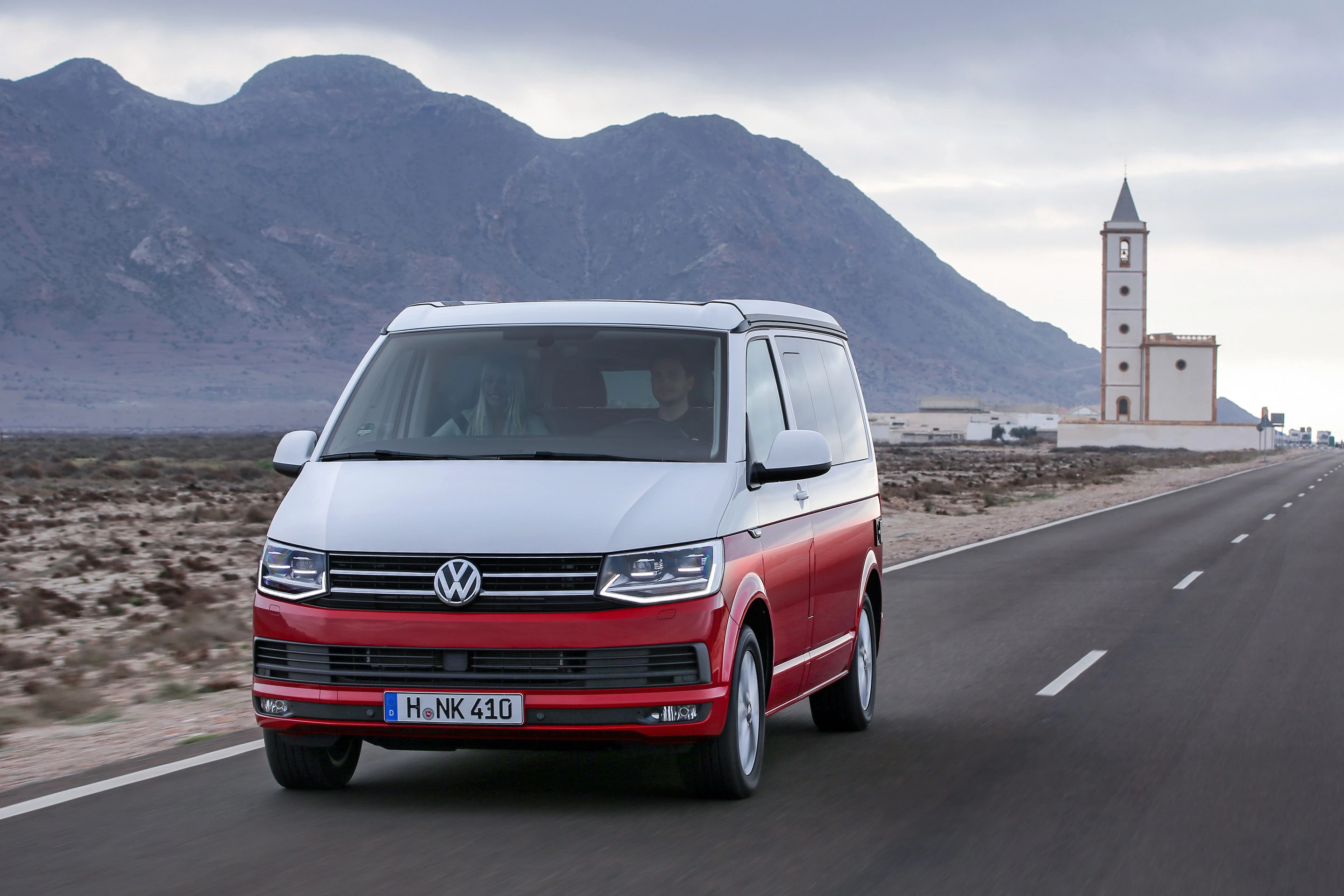 Ein weiß-roter Volkswagen Bus fährt auf einer Landstraße, im Hintergrund sind Berge und eine Kirche zu sehen.
