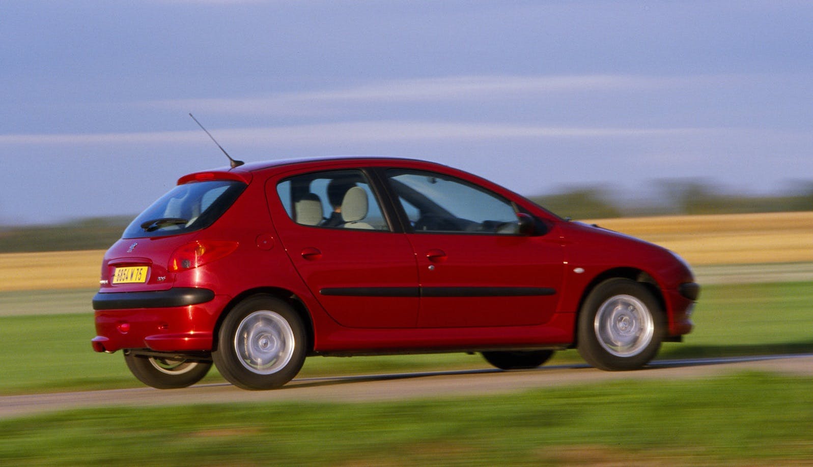 Roter Peugeot 206 fährt schnell auf einer Landstraße durch eine ländliche Gegend, unscharfer Hintergrund, sonniges Wetter.