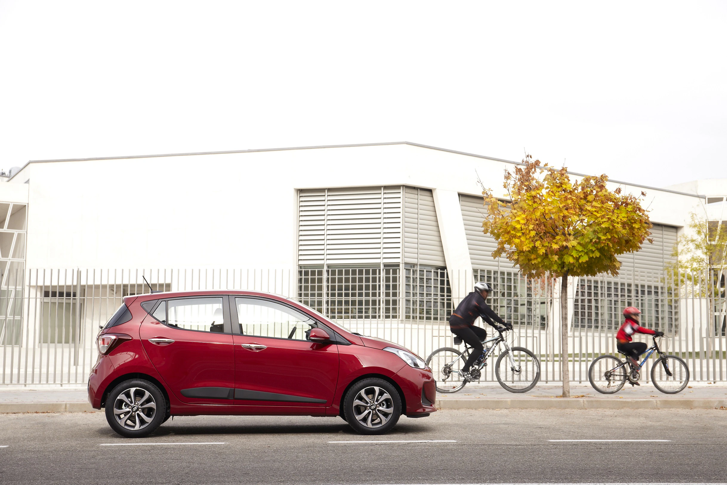 Rotes Auto parkt vor modernem Gebäude. Zwei Radfahrer, ein Erwachsener und ein Kind, fahren auf einer Straße. Herbstbaum im Hintergrund.