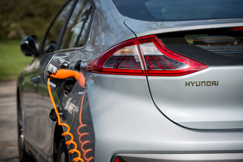 Silbernes Elektroauto der Marke Hyundai wird mit einem orangefarbenen Kabel aufgeladen, das an der linken hinteren Seite angeschlossen ist.