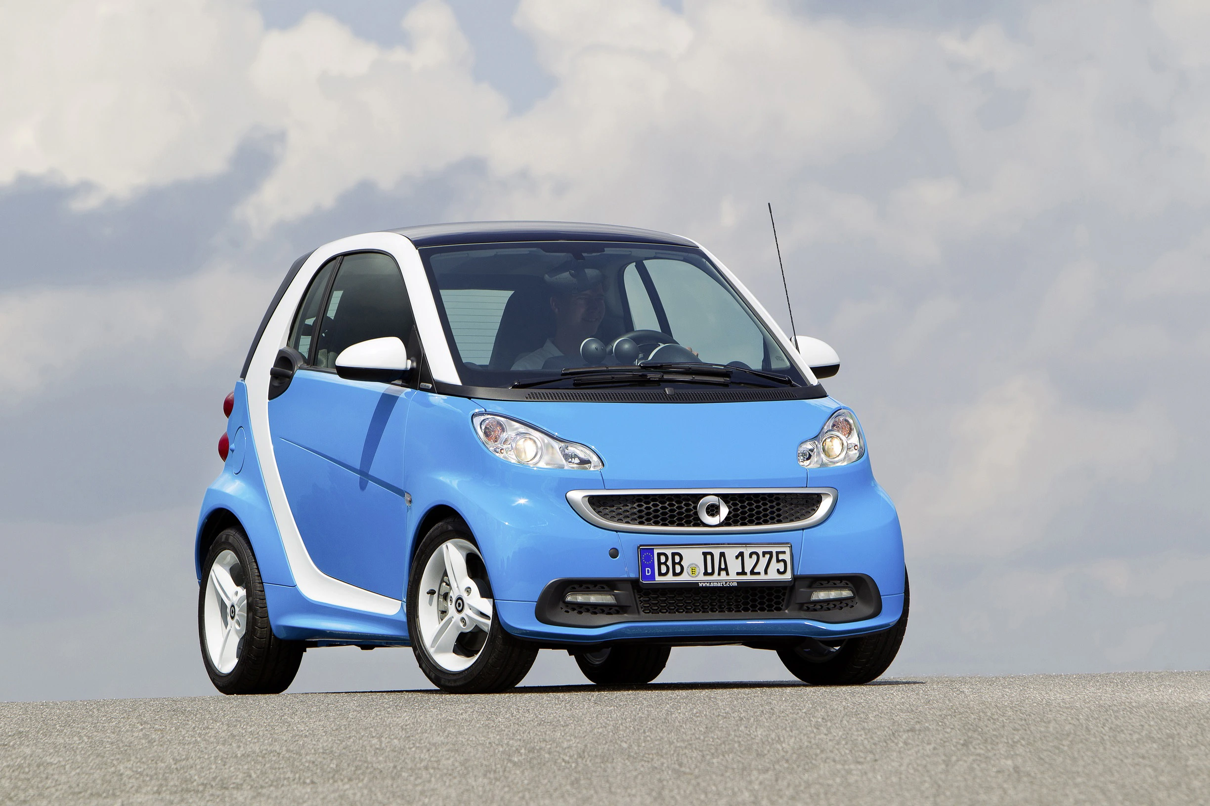 Blauer Smart Fortwo steht auf einer Straße vor bewölktem Himmel. Der Kleinwagen hat ein kompaktes Design mit weißer Akzentuierung.