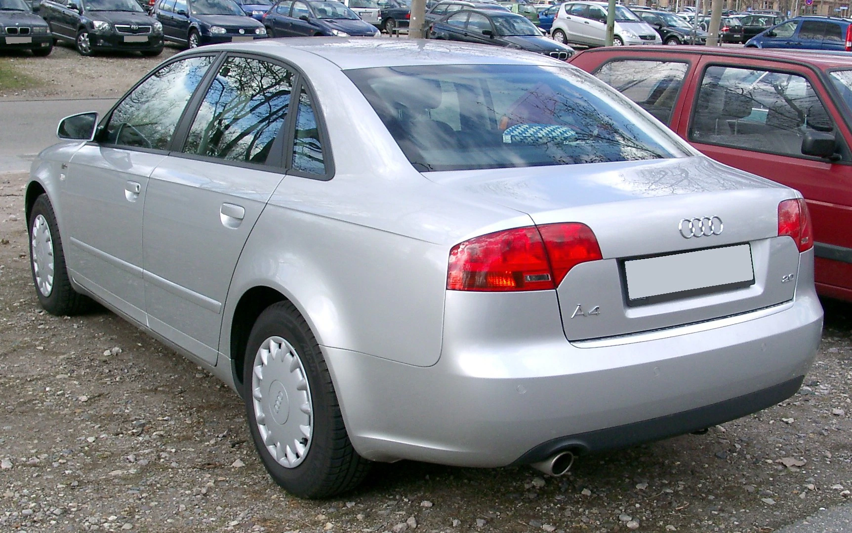 Silberner Audi A4 Limousine, seitliche Heckansicht, geparkt auf Schotterplatz, umgeben von anderen Autos.