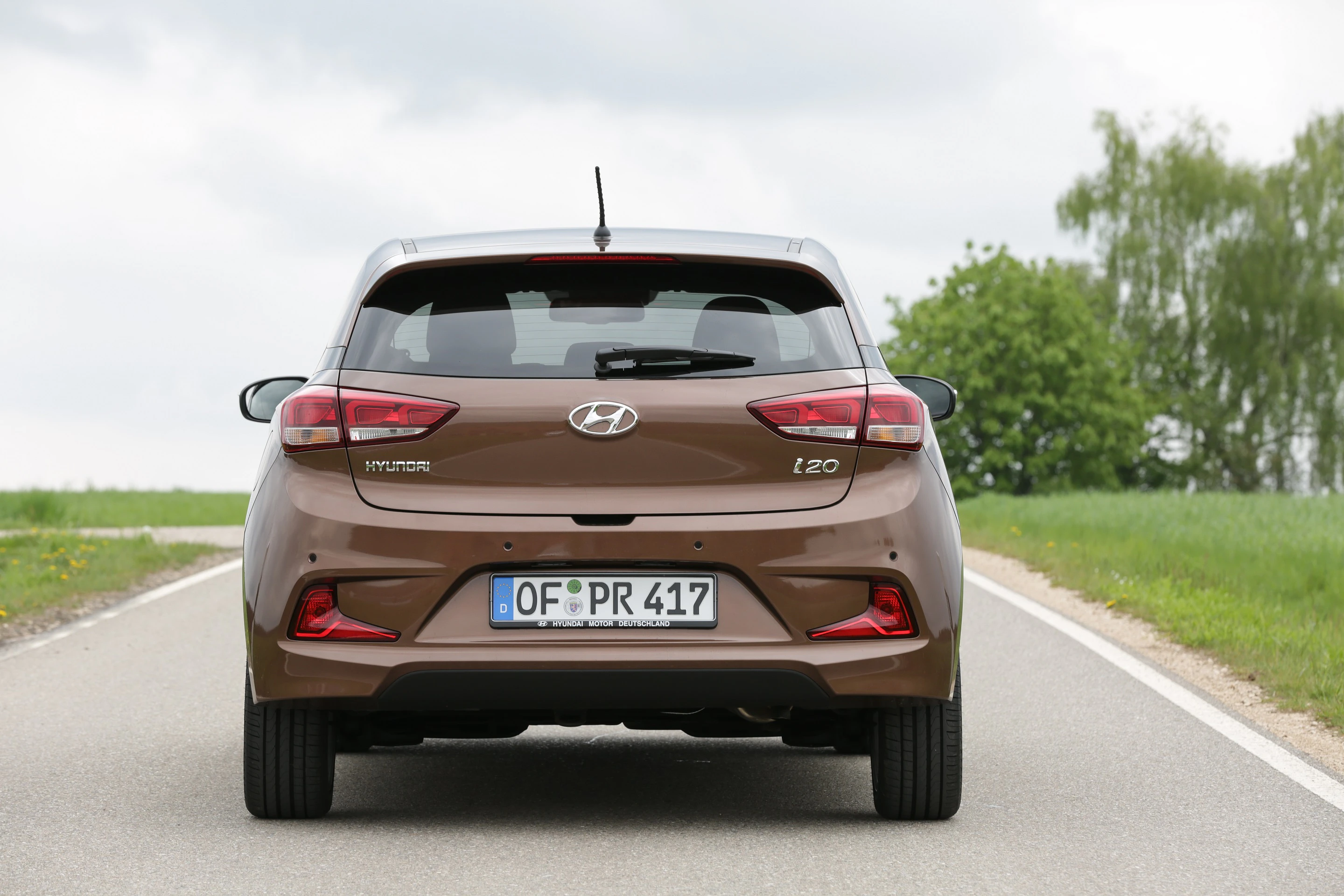 Brauner Hyundai i20 von hinten auf einer Landstraße, umgeben von grünen Feldern und Bäumen unter bewölktem Himmel.