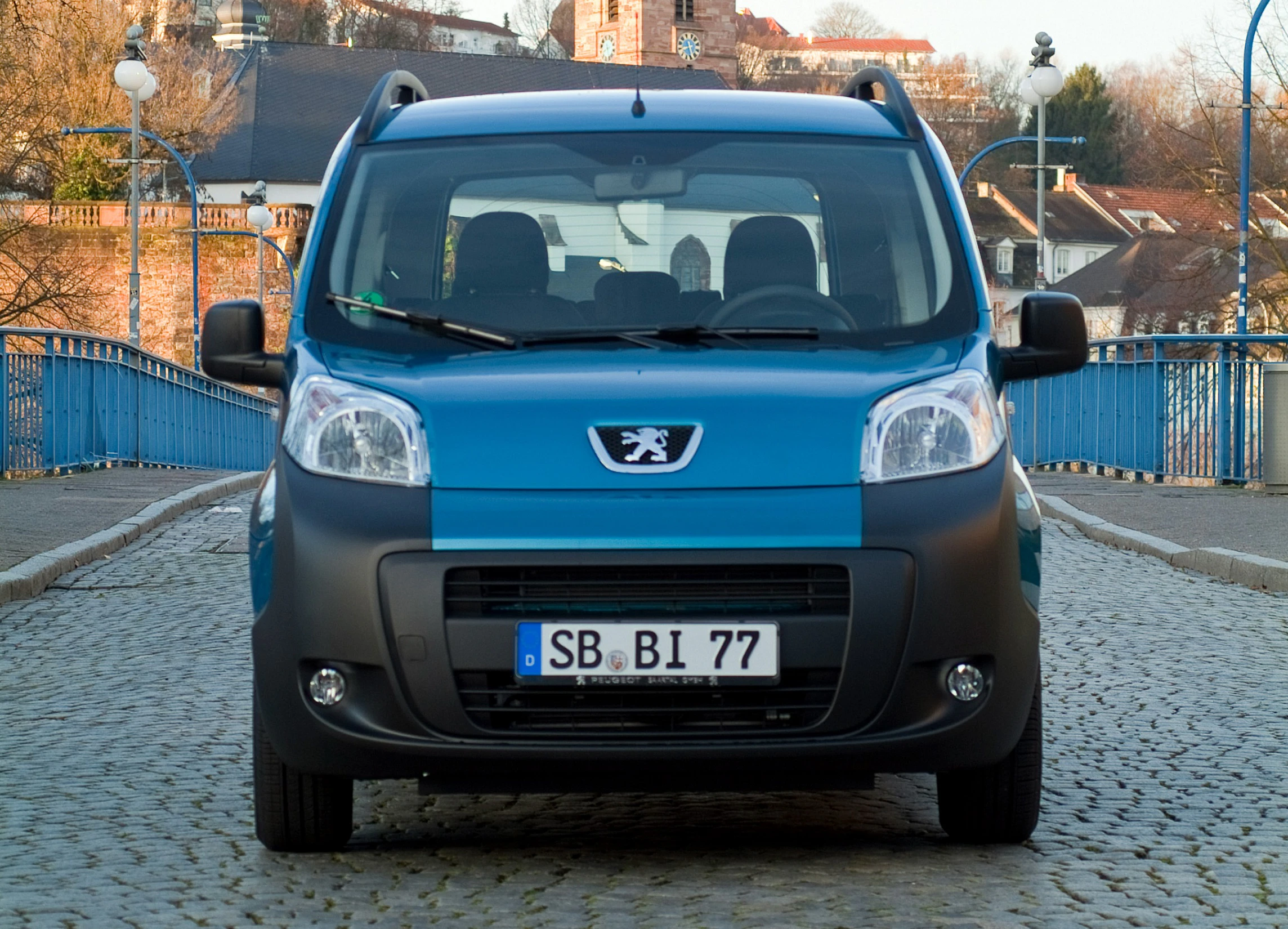 Ein blauer Peugeot Kleintransporter steht auf einer gepflasterten Brücke mit Stadtansicht im Hintergrund.