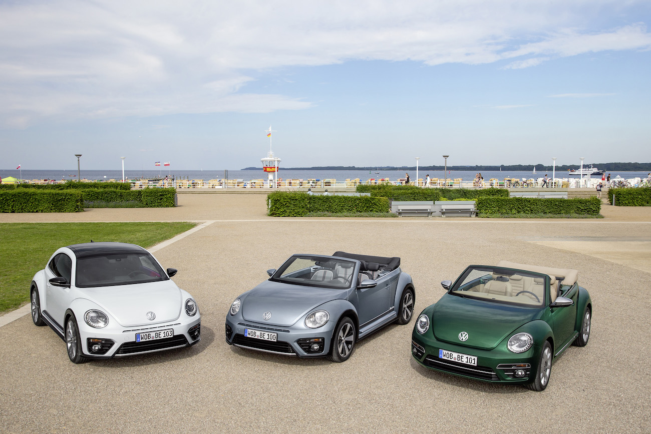 Drei VW Beetle Cabriolets in verschiedenen Farben stehen nebeneinander auf einem gepflasterten Platz mit Strand und Meer im Hintergrund.