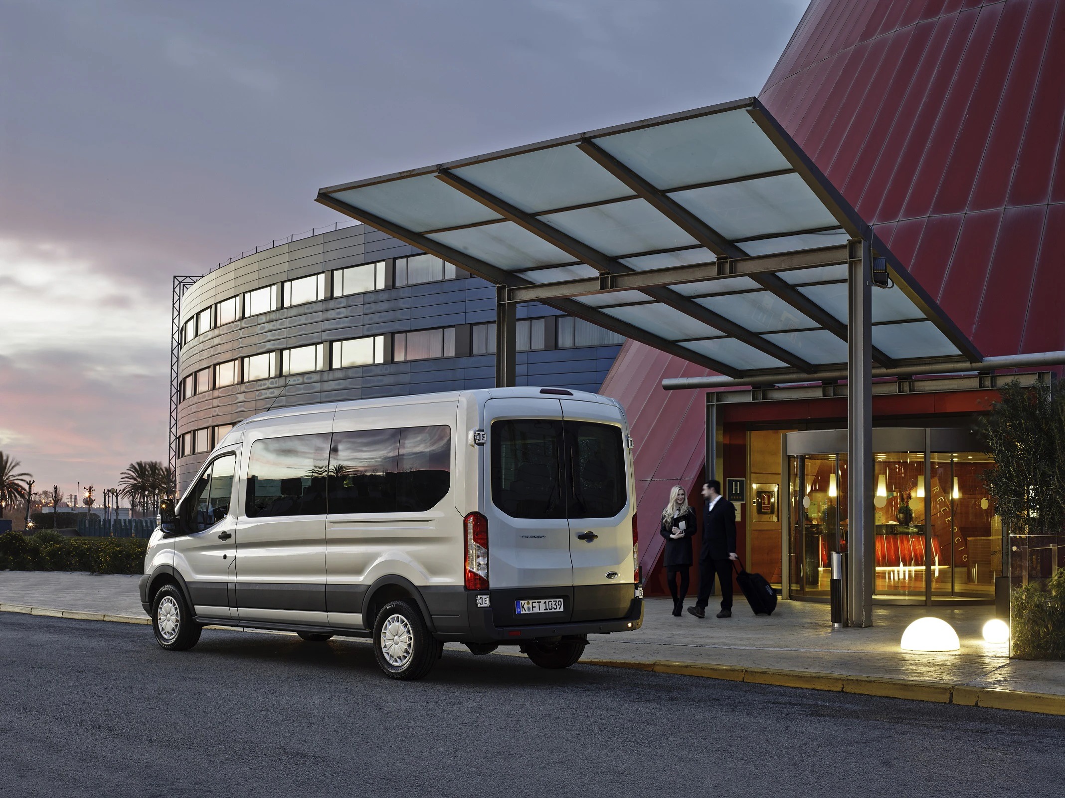 Silberner Van vor einem modernen Gebäude mit Glasdach, zwei Personen mit Koffern sind am Eingang, Sonnenuntergang im Hintergrund.
