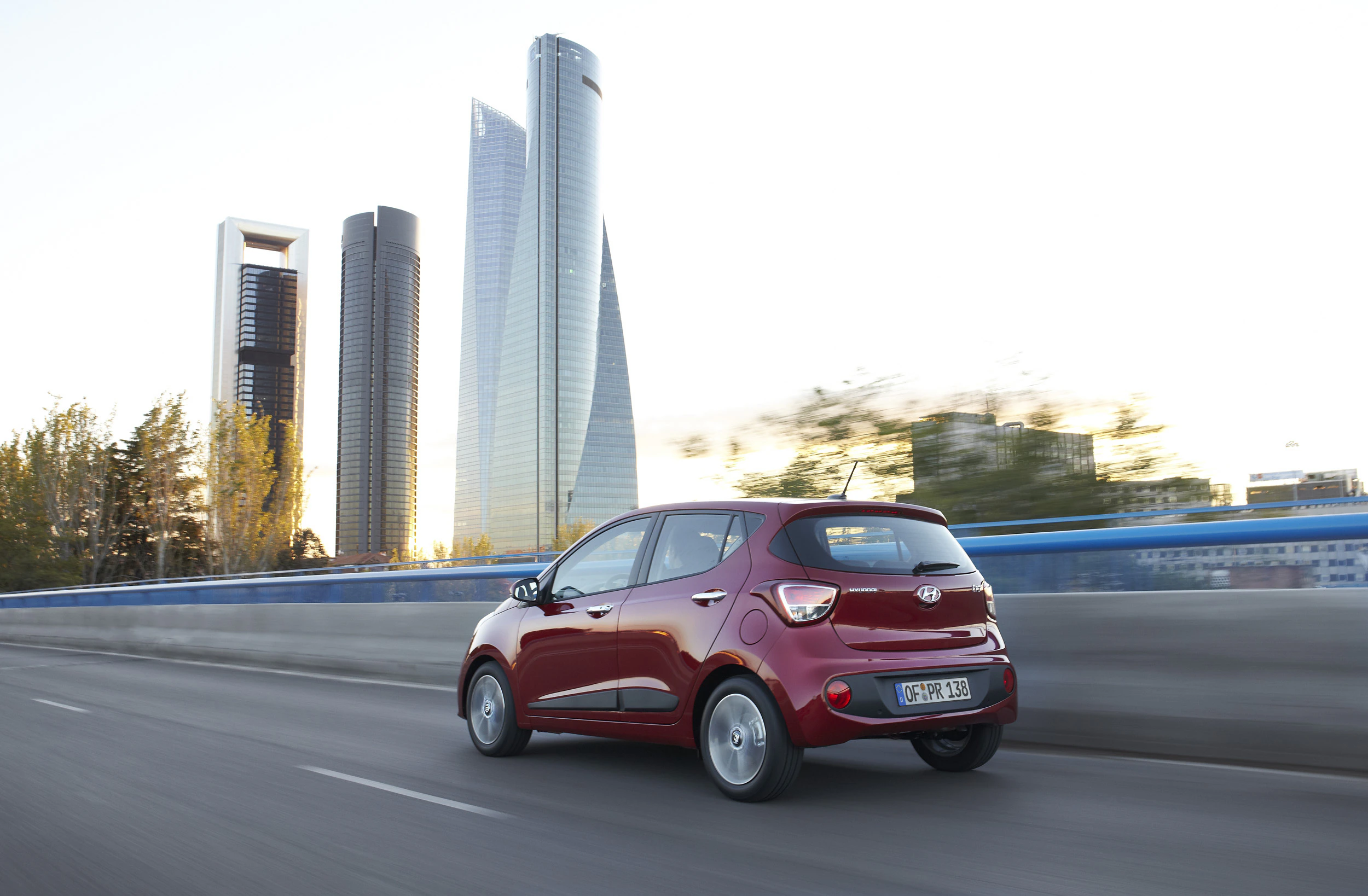 Ein rotes Auto fährt auf einer Autobahn, im Hintergrund sind moderne Wolkenkratzer unter klarem Himmel in einer Stadt zu sehen.