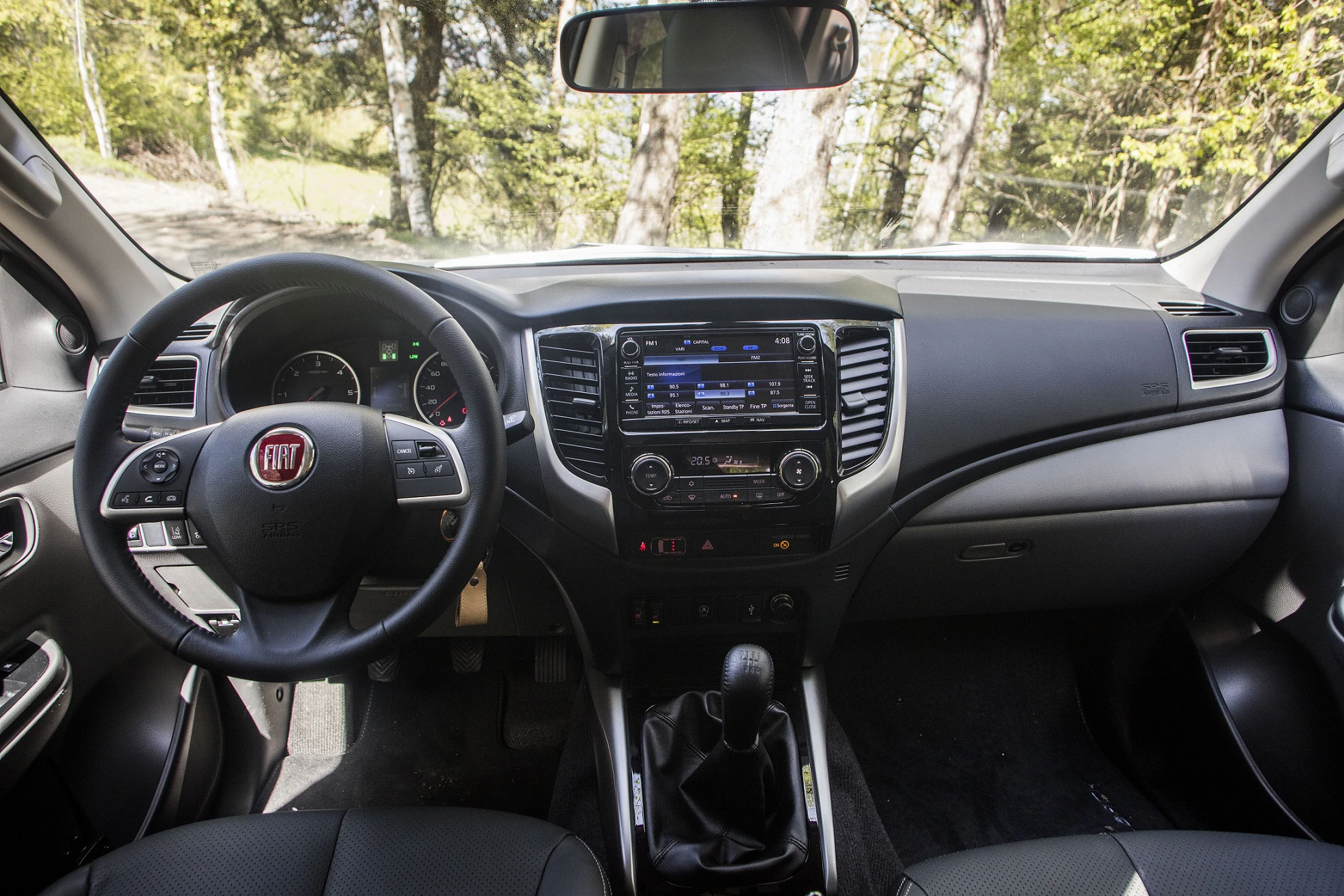 Innenraum eines Autos mit grauem Armaturenbrett, Lenkrad links, zentralem Touchscreen, Klimaanlage, Schaltknüppel und Außensicht auf Bäume.