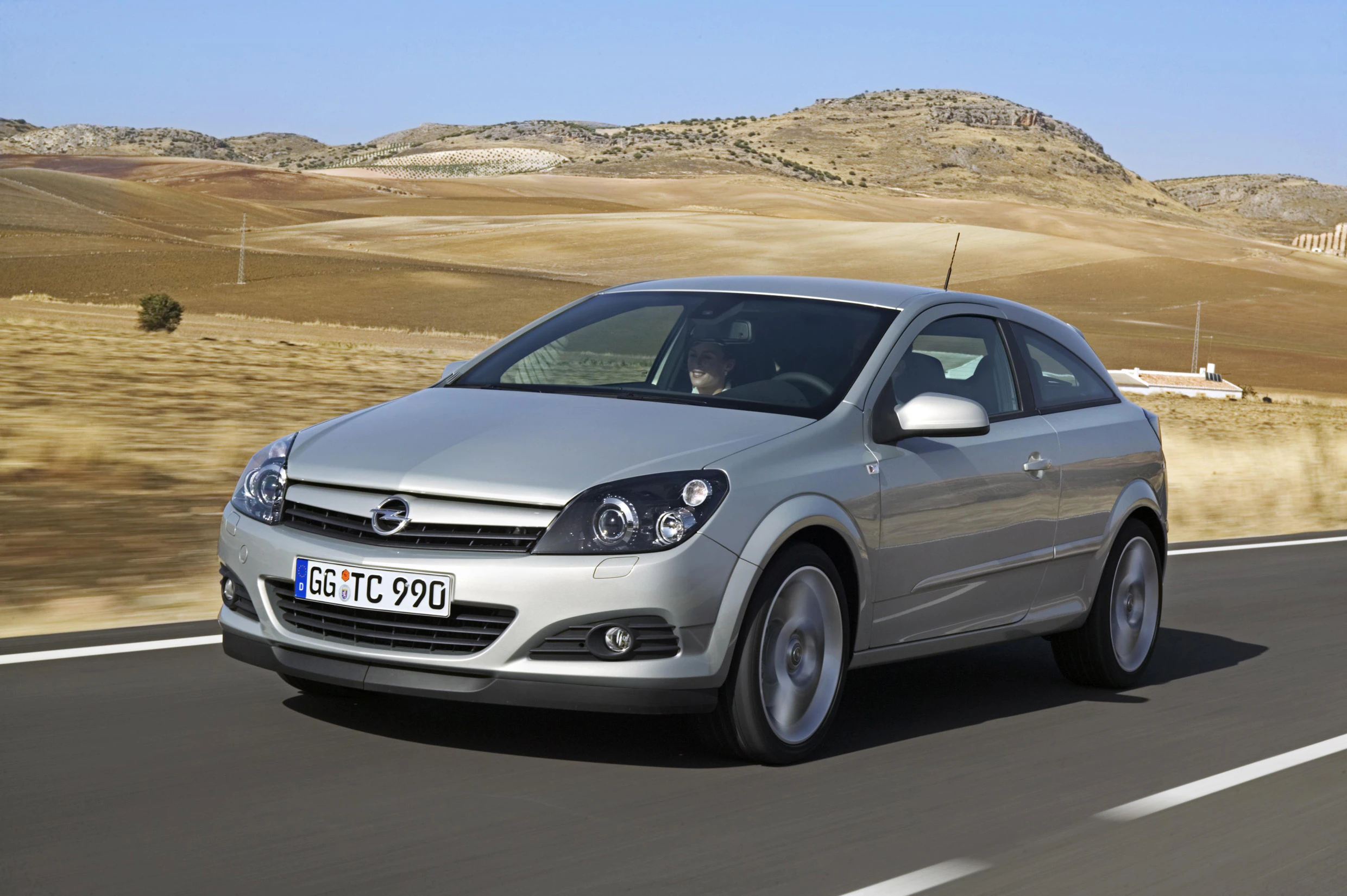 Silberner Opel Astra fährt auf Landstraße durch trockene Landschaft unter klarem Himmel.