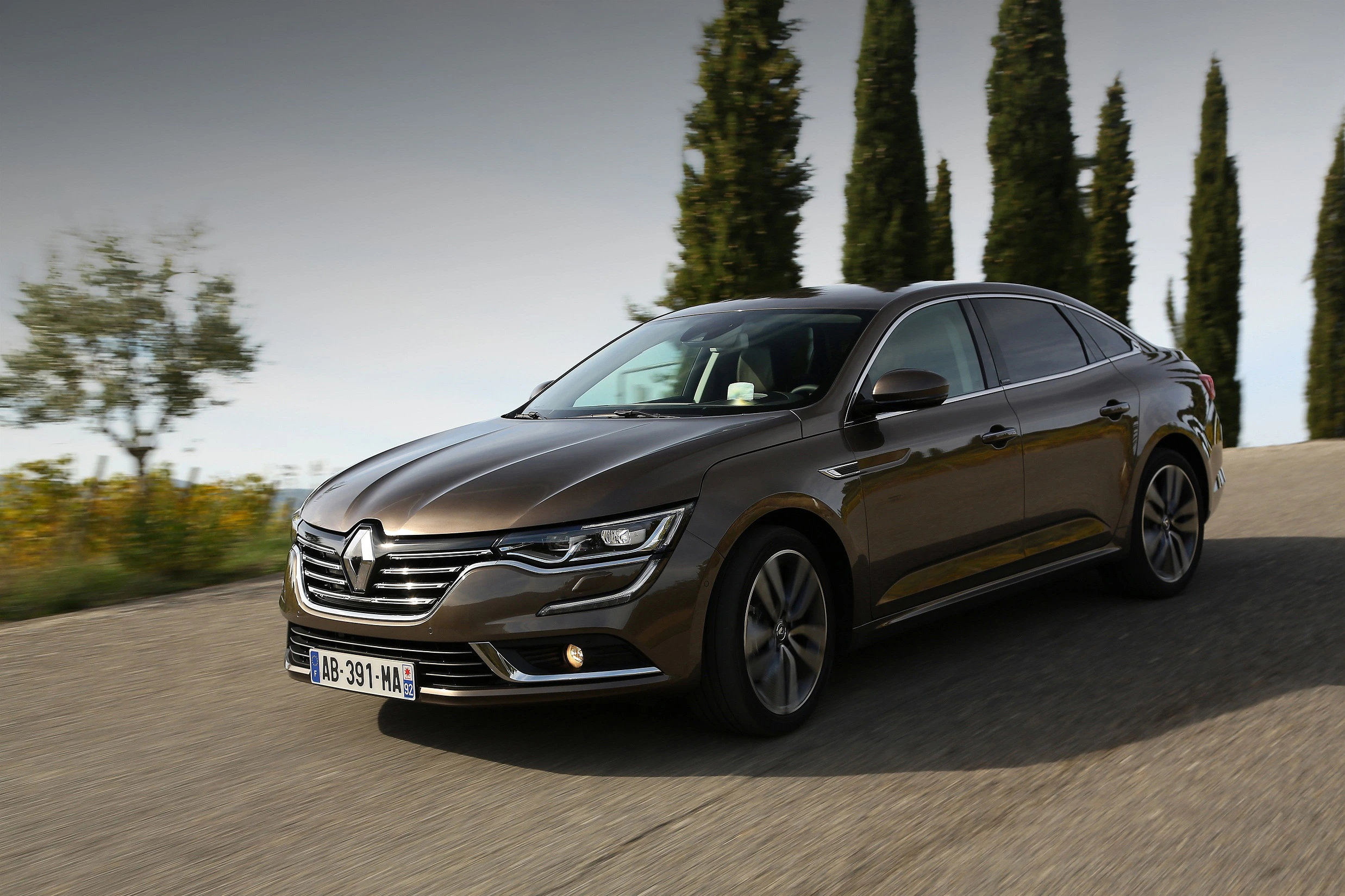 Brauner Renault Talisman fährt auf einer Landstraße, flankiert von Zypressen und blühenden Wiesen, unter einem bewölkten Himmel.