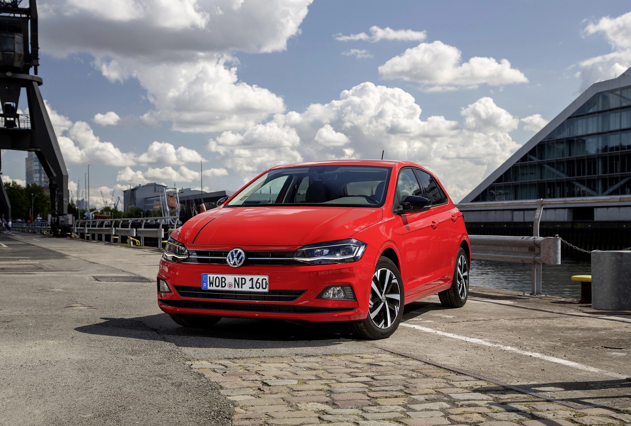 Roter Volkswagen Polo parkt auf einem Hafengelände bei sonnigem Wetter. Im Hintergrund sind moderne Gebäude und ein Kran zu sehen.