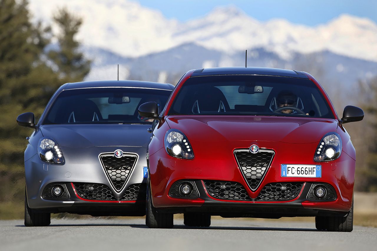 Zwei Alfa Romeo Giulias, eine rot, eine grau, fahren auf einer Straße vor Bergkulisse.