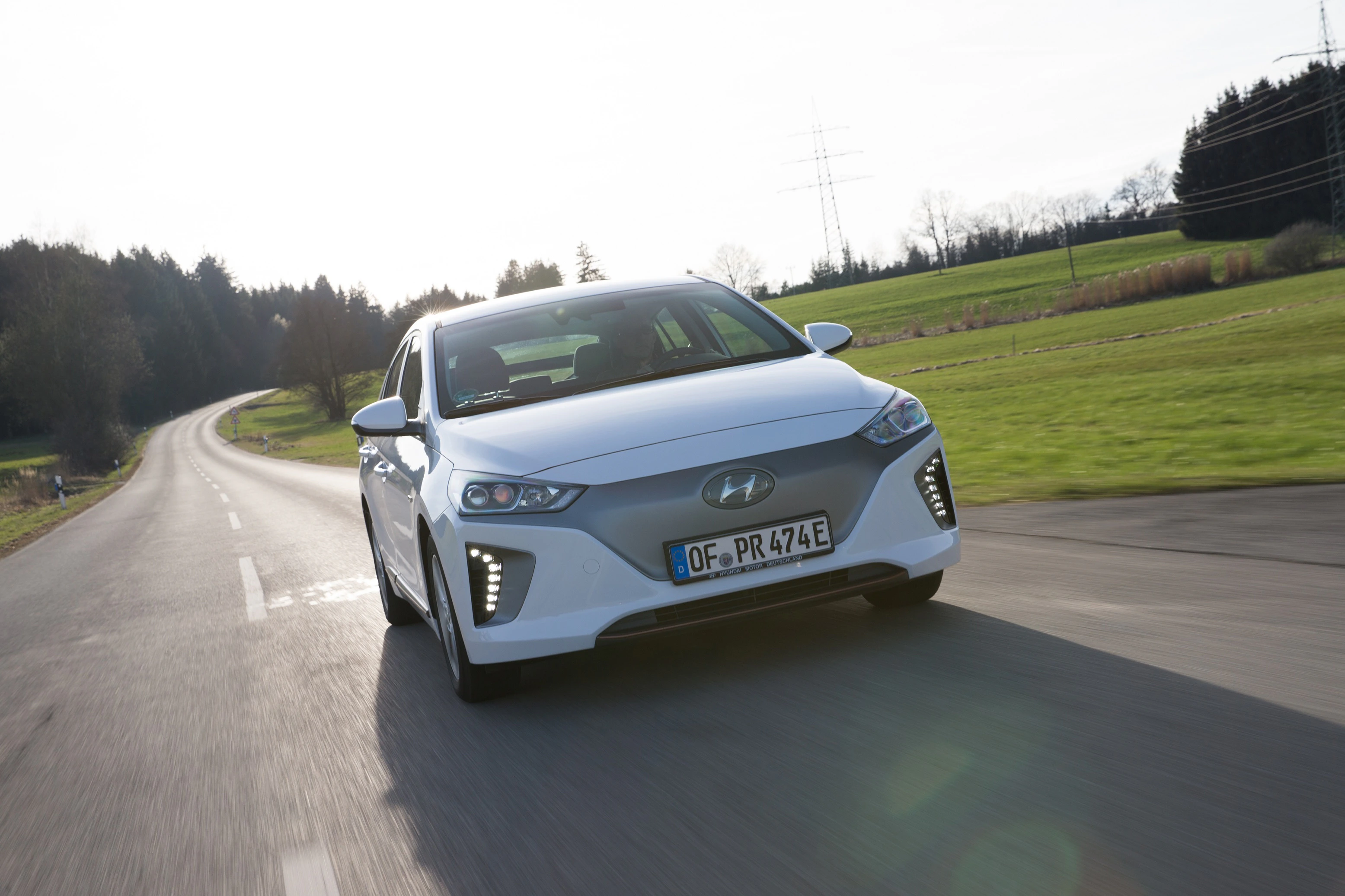Weißes Elektroauto von Hyundai fährt auf einer Landstraße durch eine grüne Landschaft an einem sonnigen Tag.