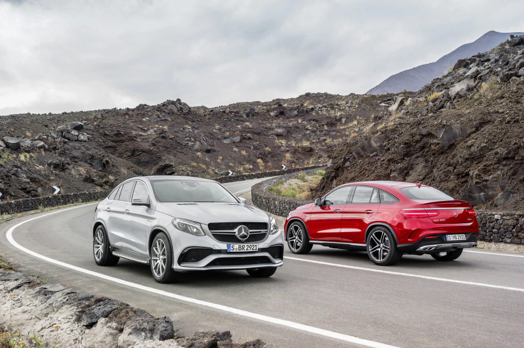 Zwei Mercedes-Benz SUVs, silber und rot, fahren auf einer kurvigen Bergstraße mit felsiger Landschaft im Hintergrund unter bewölktem Himmel.