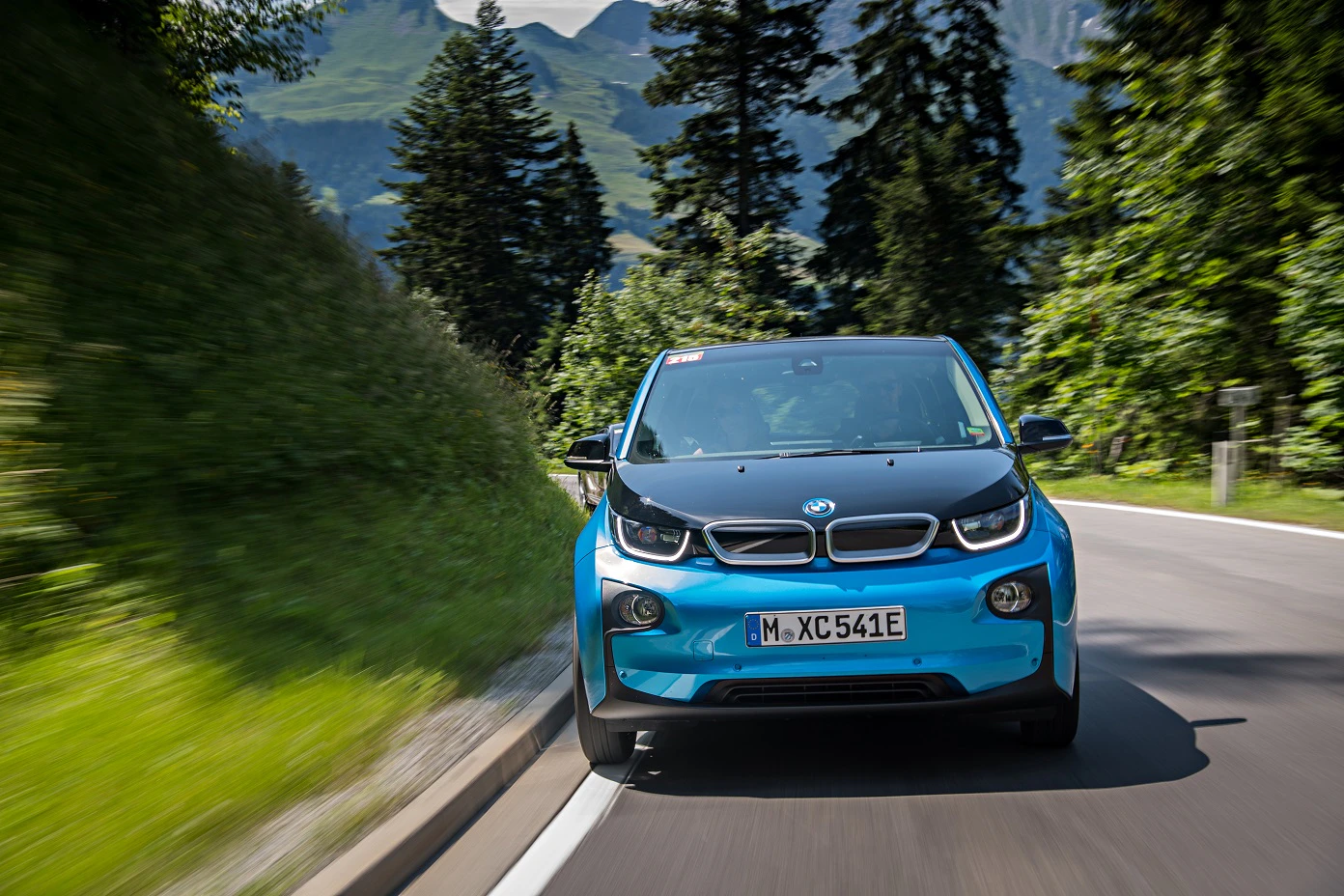 Blauer BMW i3 fährt auf einer Straße durch eine bewaldete Berglandschaft an einem sonnigen Tag.