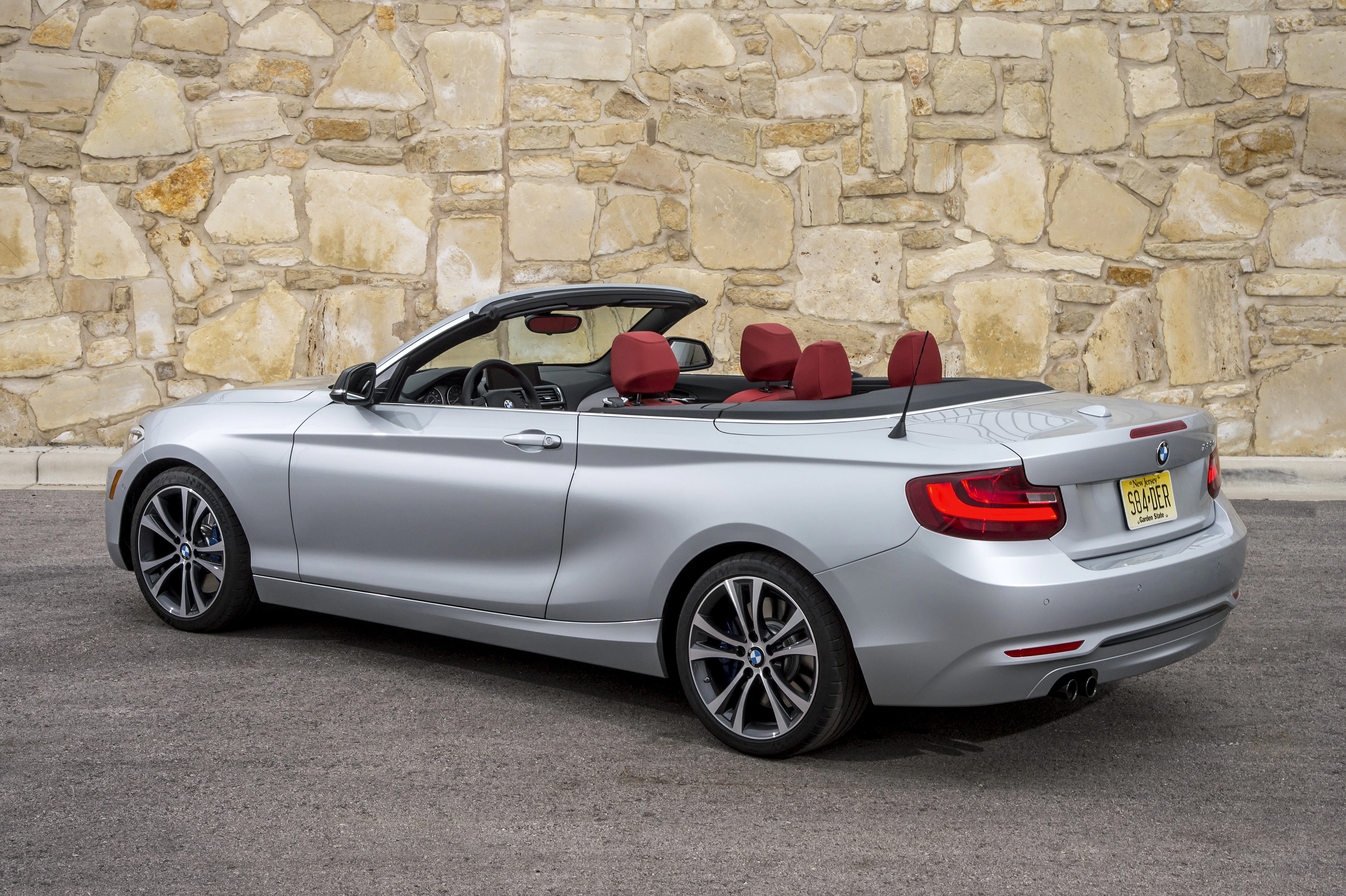 Silbernes Cabriolet mit roter Innenausstattung vor einer Steinmauer, aus der Hecksicht aufgenommen. Fahrzeug hat ein offenes Verdeck.