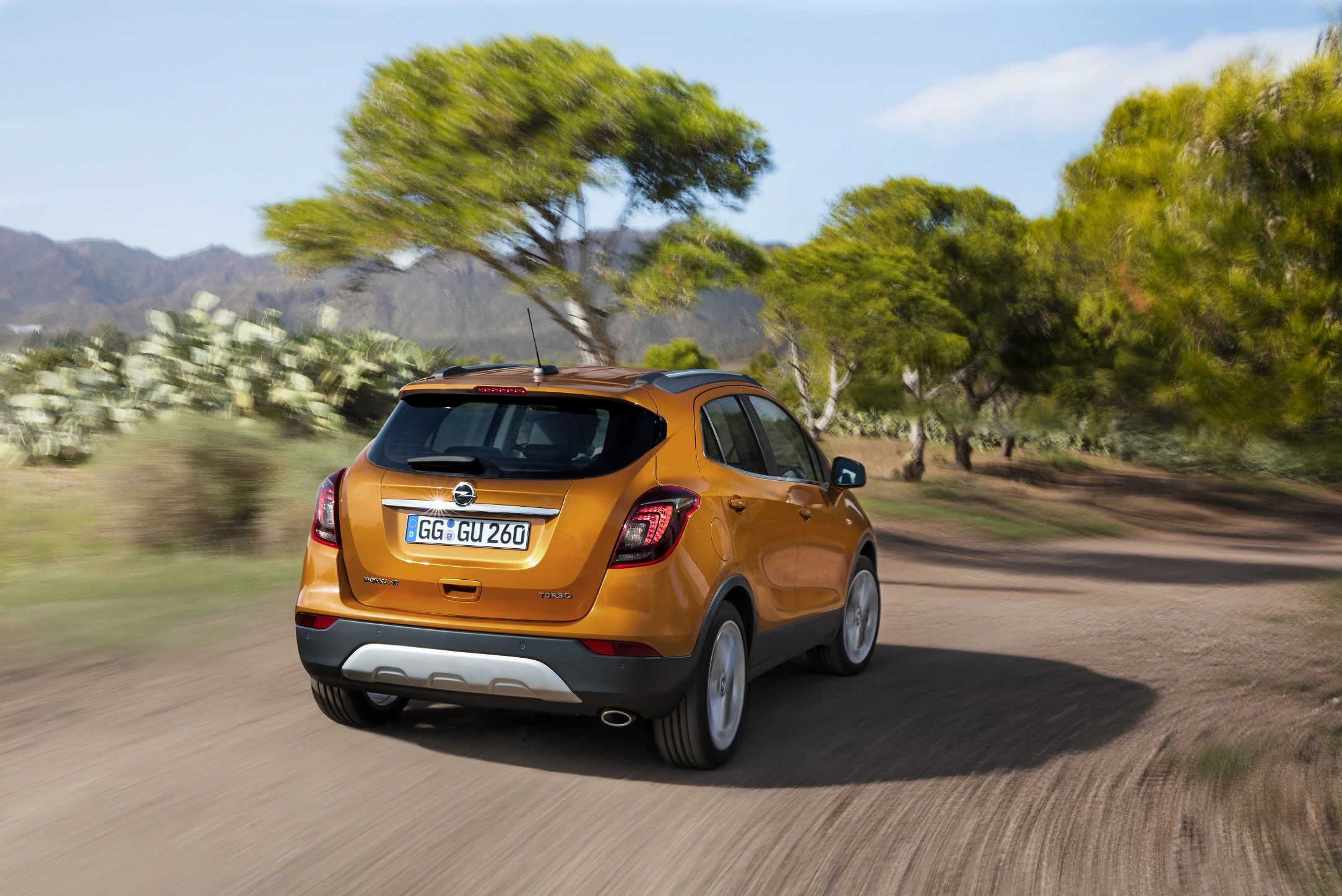 Orangefarbener SUV fährt auf einem staubigen Weg durch eine landschaftlich reizvolle Umgebung mit Bäumen und Bergen im Hintergrund.