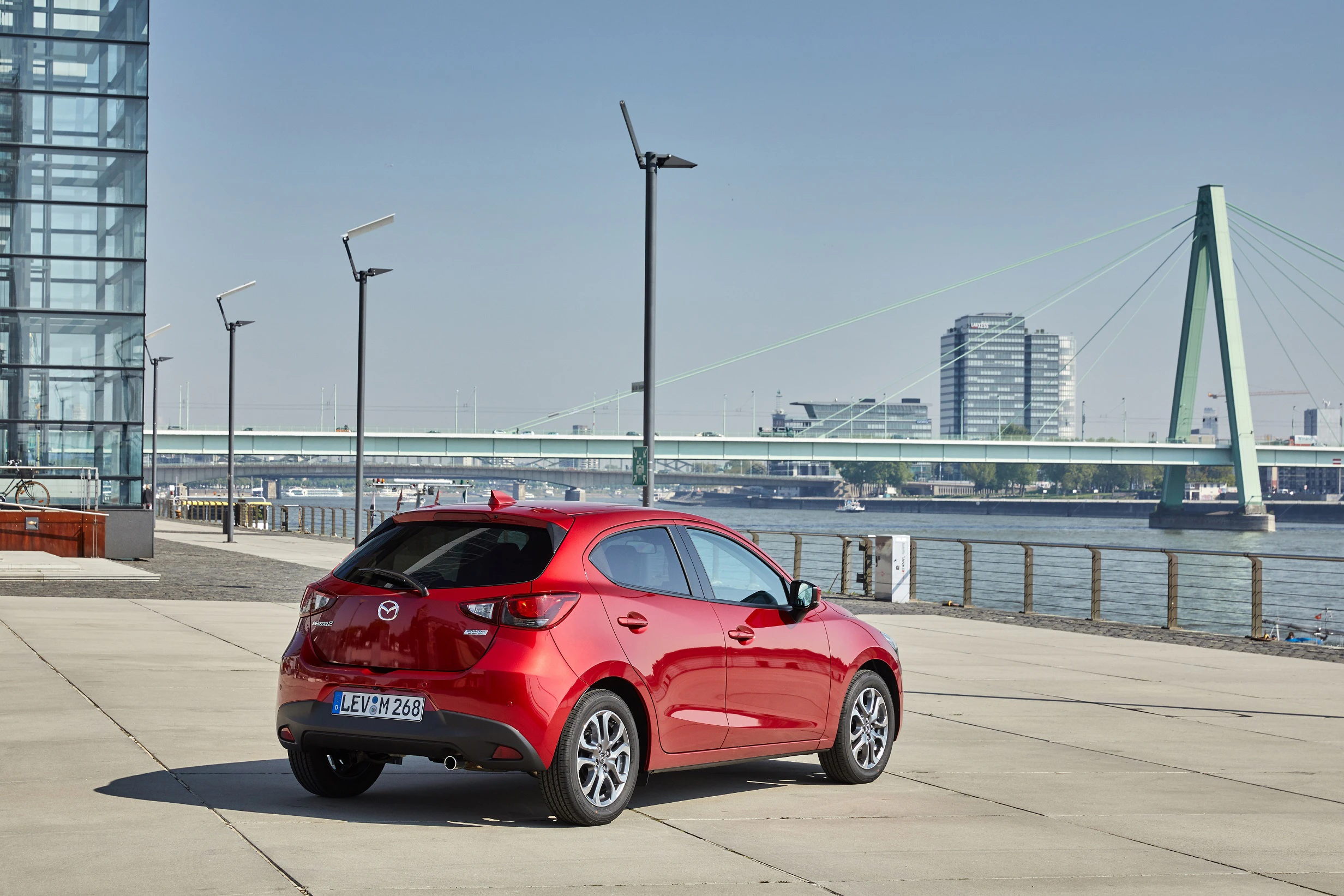 Rotes Auto auf einem gepflasterten Platz am Flussufer, moderne Gebäude und eine Brücke im Hintergrund. Klarer, sonniger Himmel.