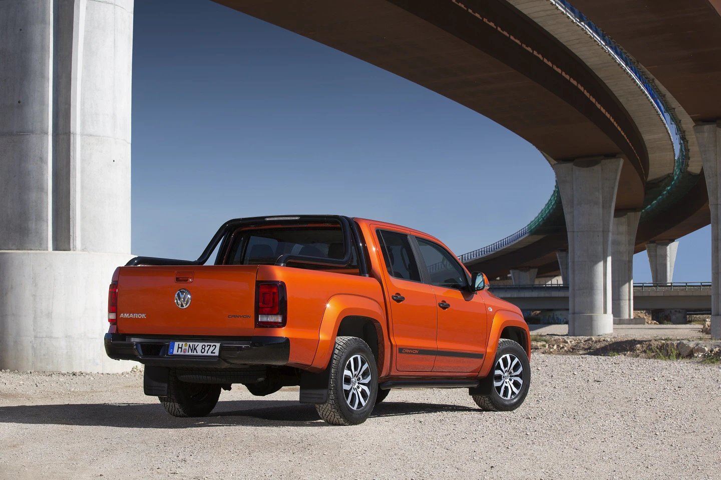 Oranger Pickup-Truck parkt unter weitläufigen Autobahnbrücken auf einem Schotterplatz. Klarer Himmel im Hintergrund.