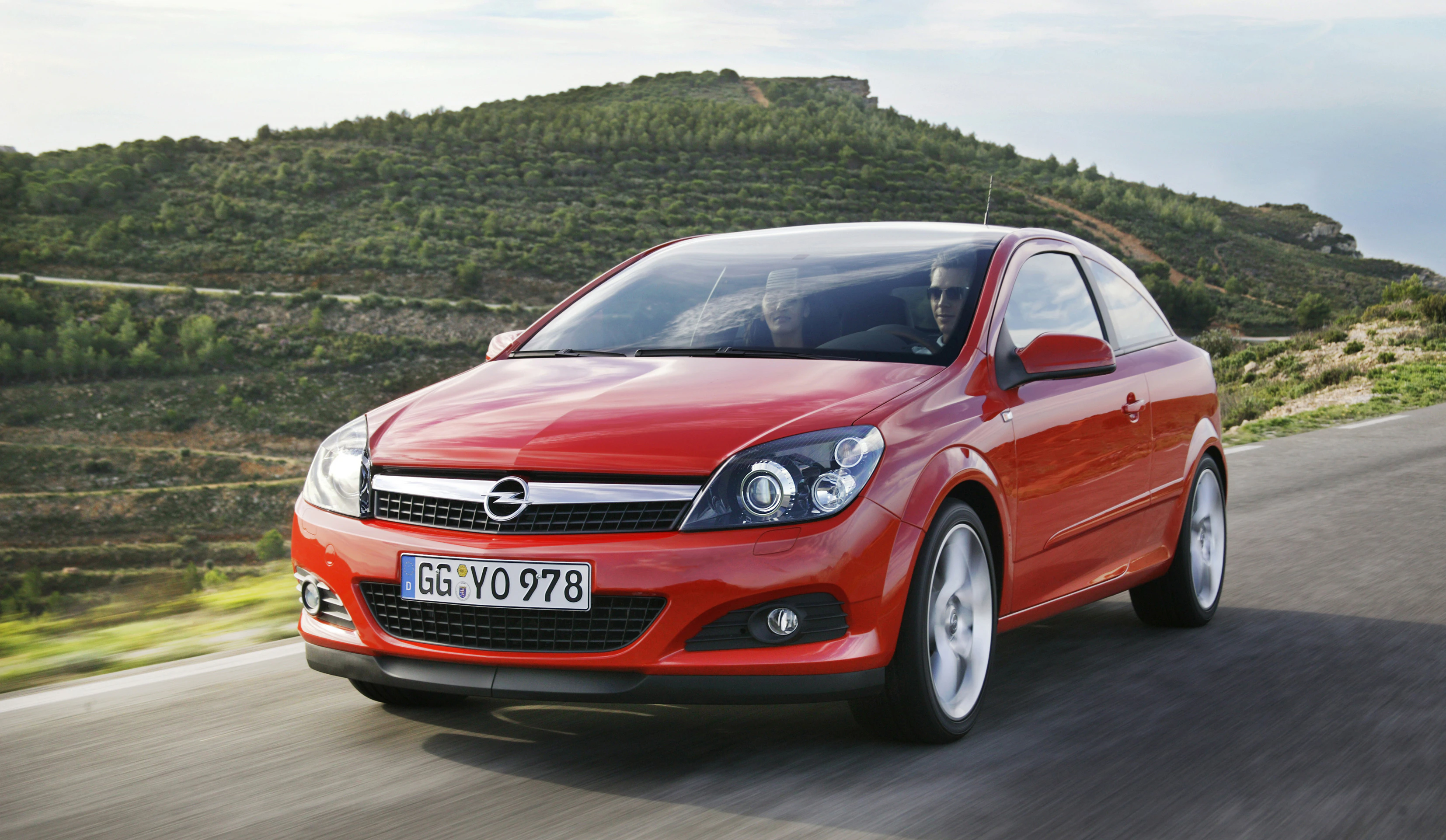 Roter Opel Astra Cabrio fährt auf einer Landstraße mit grünen Hügeln im Hintergrund. Klarer Himmel, dynamische Darstellung der Bewegung.