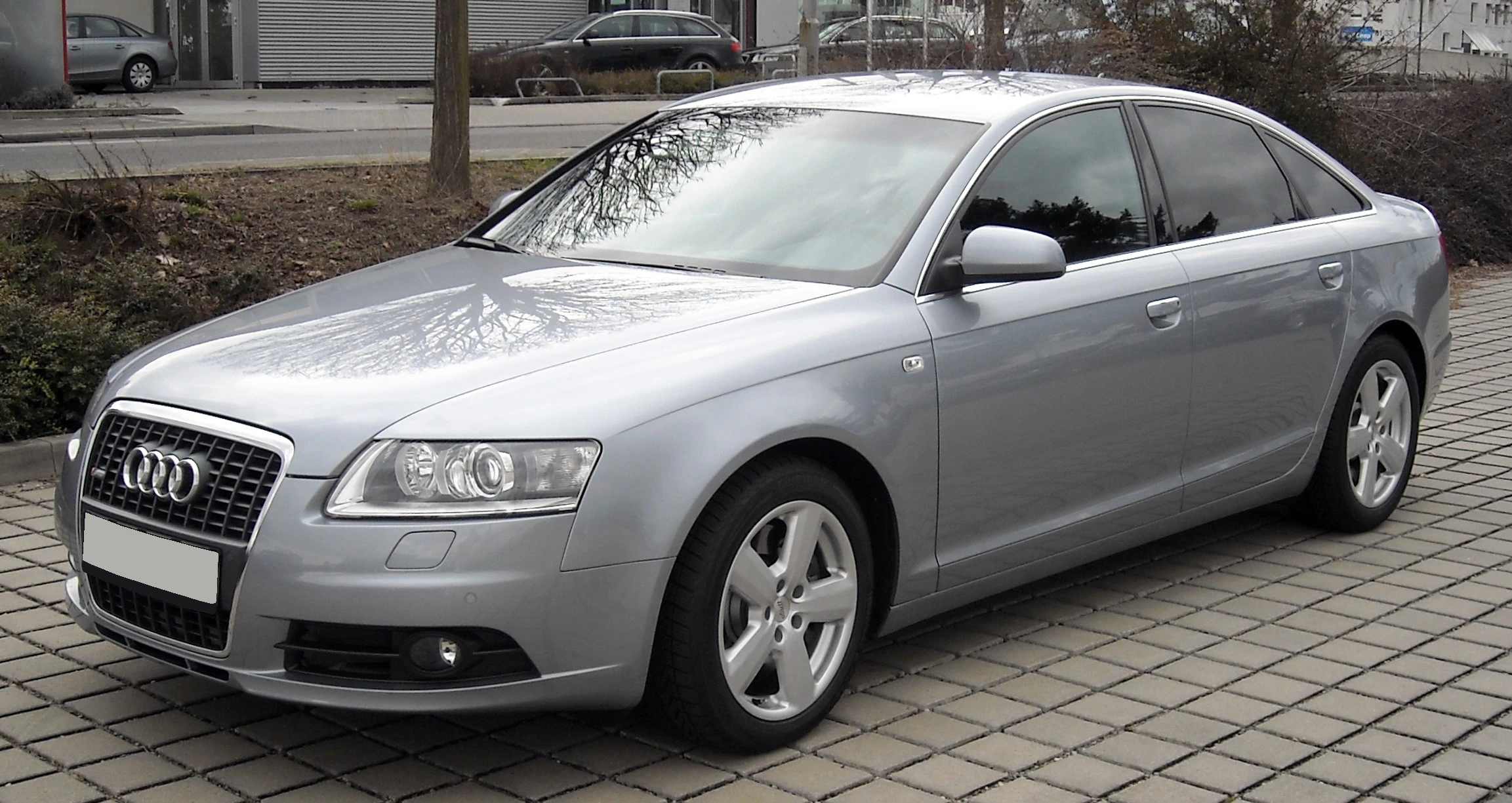 Silberner Audi A6 Limousine auf gepflastertem Parkplatz, seitliche Ansicht, vor städtischem Hintergrund mit Bäumen und Gebäuden.