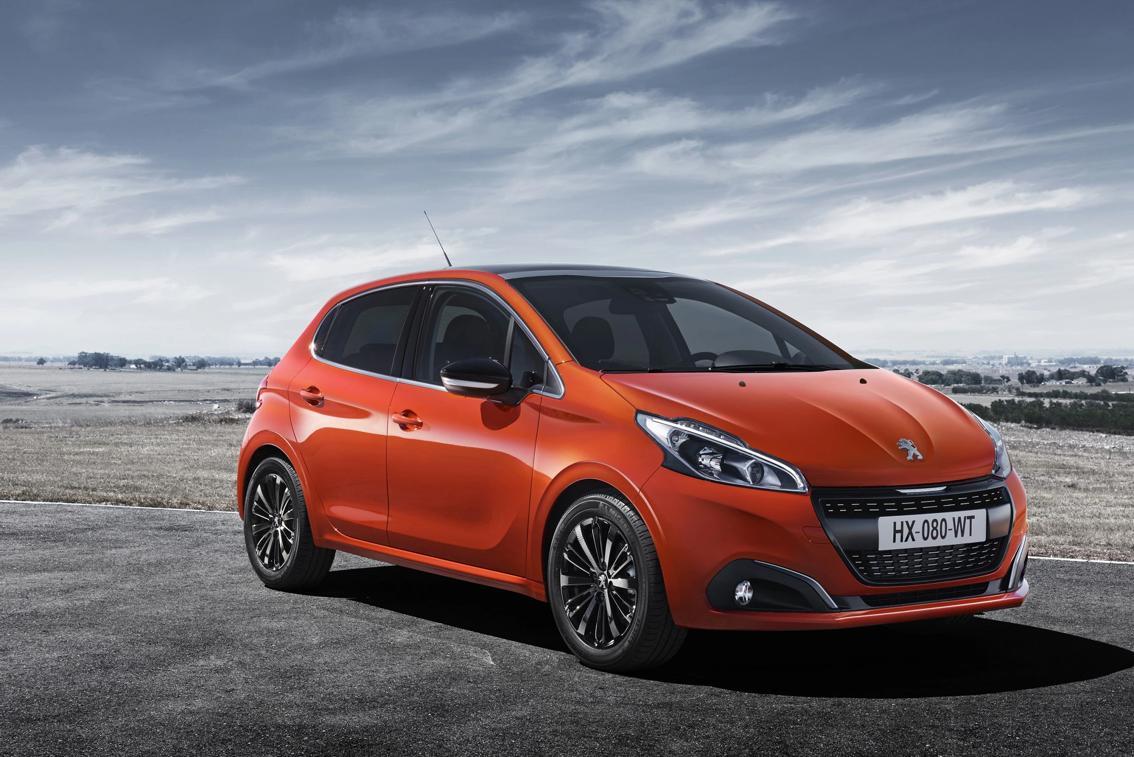 Orangefarbener Peugeot auf einer Landstraße mit wolkigem Himmel im Hintergrund.