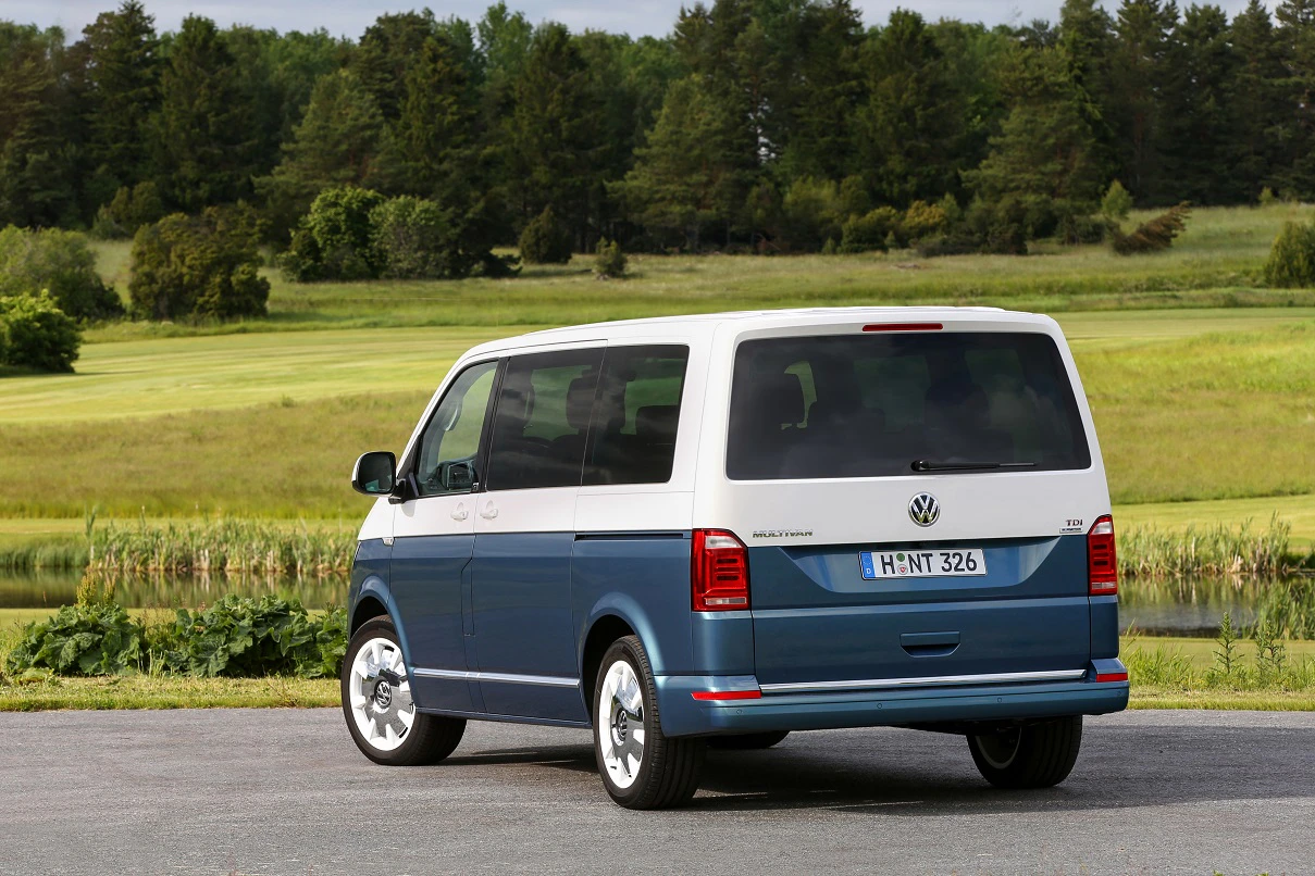 Ein zweifarbiger Volkswagen-Bus steht auf einem Parkplatz vor einer grünen Landschaft mit Bäumen.