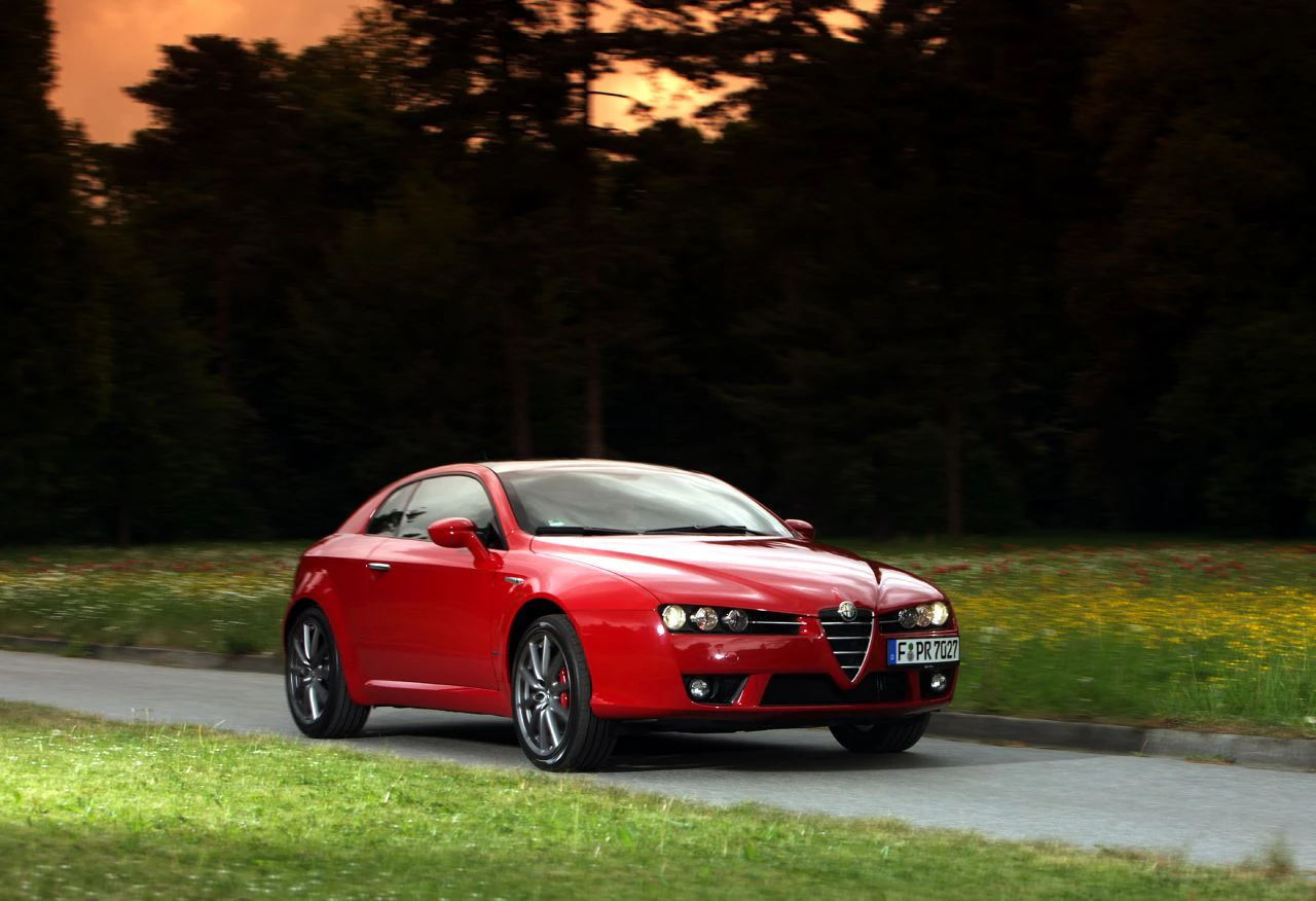 Roter Sportwagen, Alfa Romeo, fährt auf einer Straße durch eine grüne Landschaft bei Sonnenuntergang. Bäume im Hintergrund.