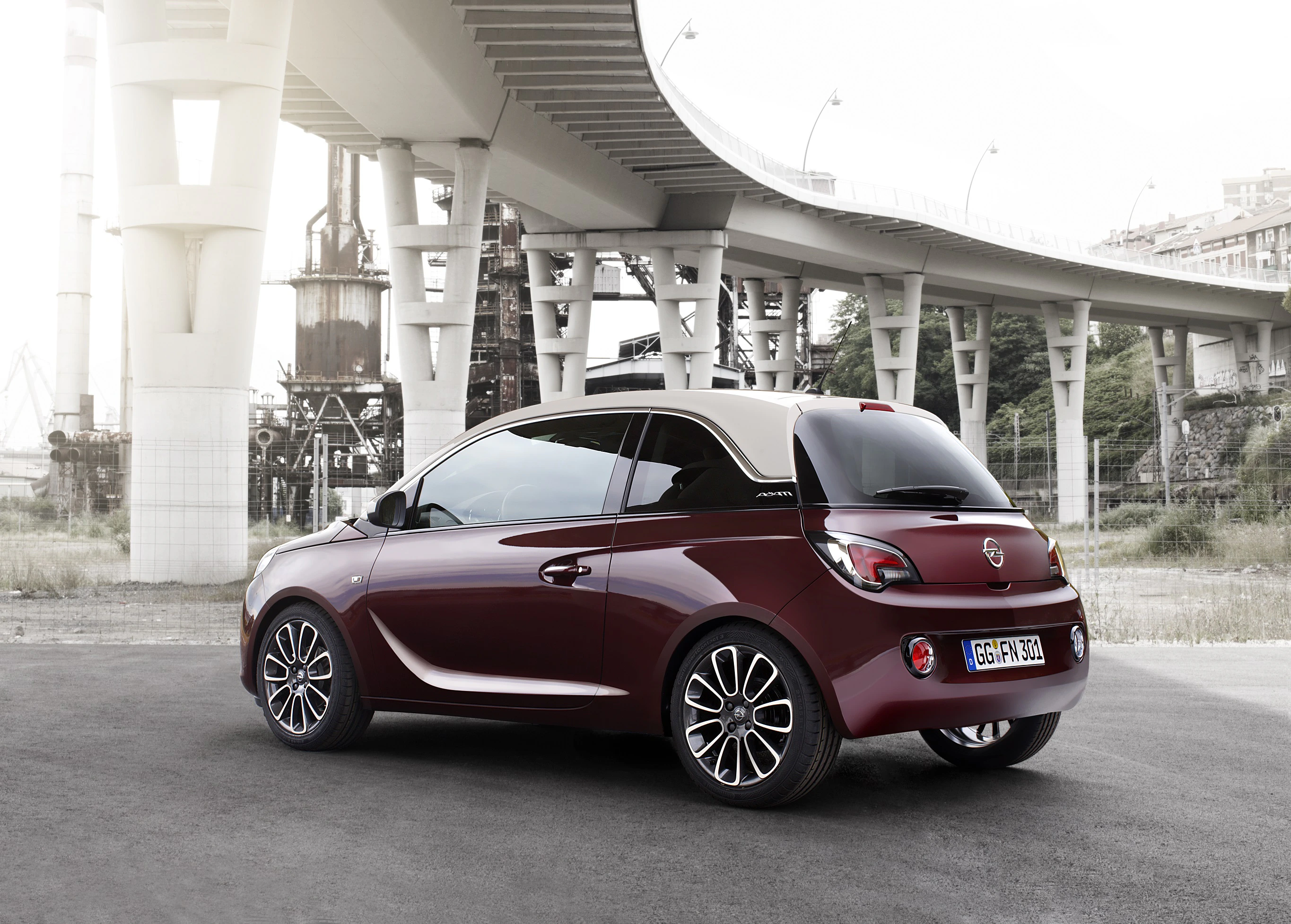 Roter Opel Adam steht auf Asphalt vor moderner Brücke. Urbaner Hintergrund mit Industrieelementen. Türlichter sind eingeschaltet.