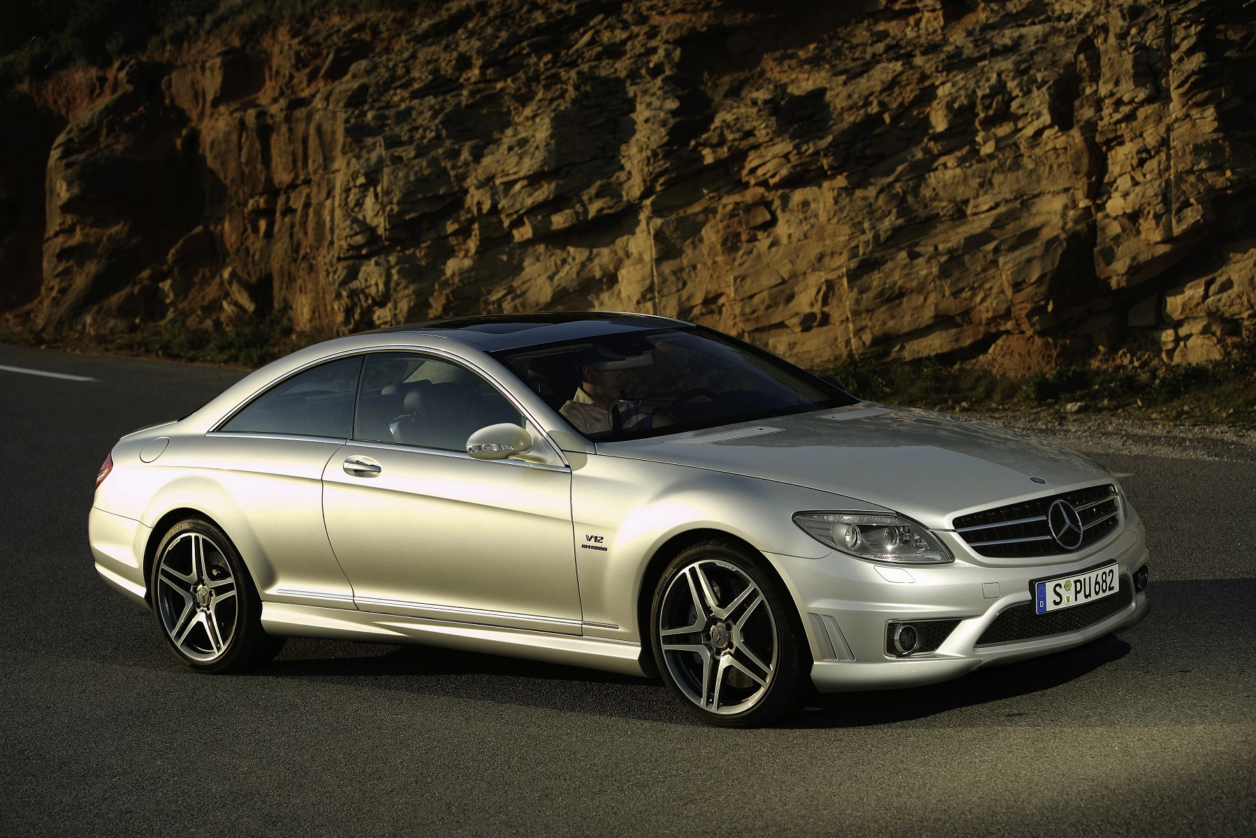 Silberner Mercedes-Benz Coupé fährt auf einer Straße neben einer Felswand, bei Sonnenuntergang.
