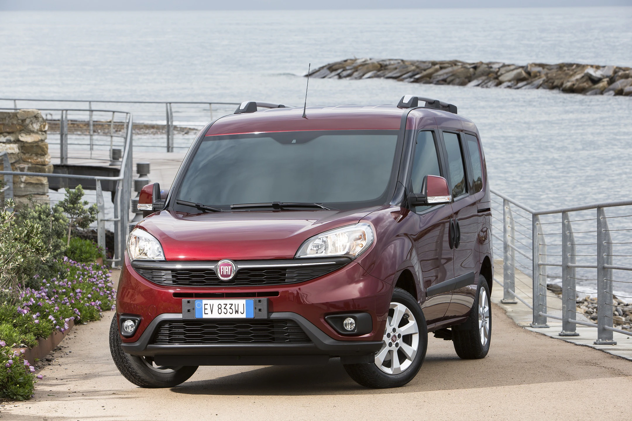 Roter Fiat Doblo parkt auf einem Weg am Meer, im Hintergrund sind Felsen und das Wasser zu sehen.