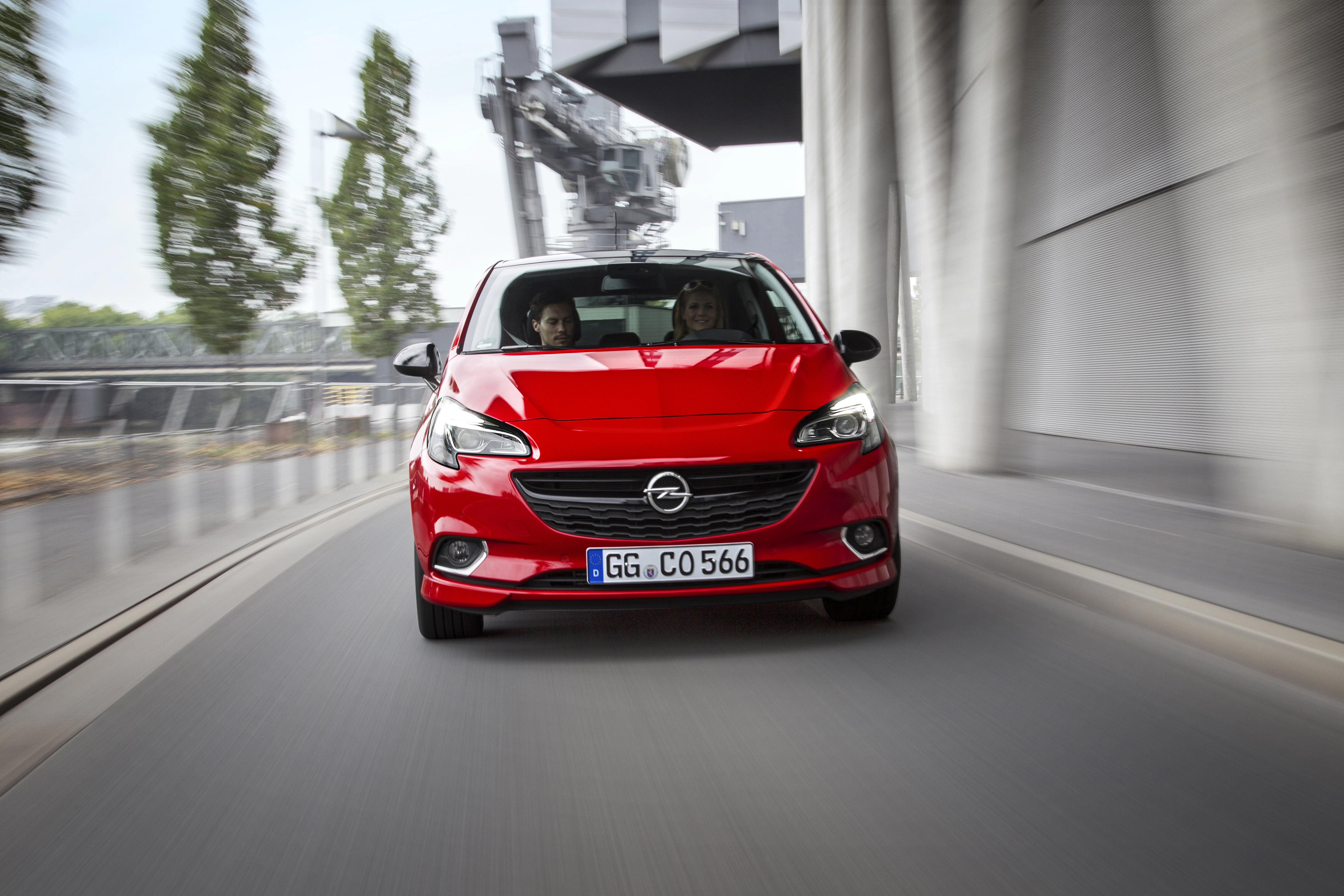 Roter Opel Corsa fährt auf einer städtischen Straße. Zwei Personen sitzen im Auto. Moderne Architektur und Bäume im Hintergrund.