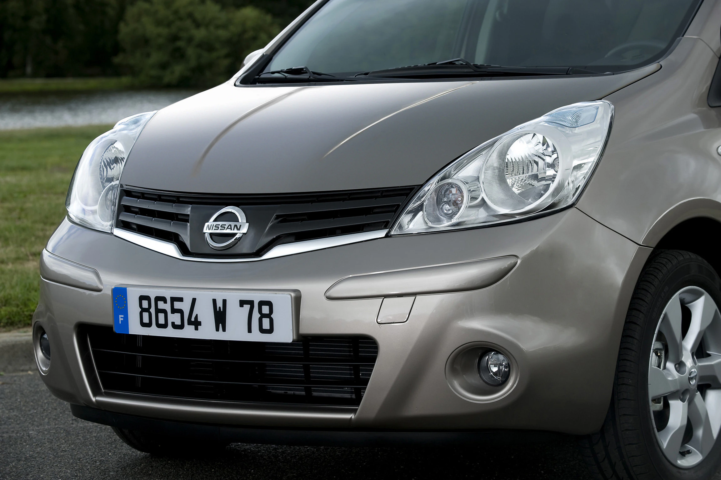 Nahaufnahme eines silbernen Nissan-Autos mit französischem Nummernschild, auf einer Straße nahe einem See im Hintergrund.