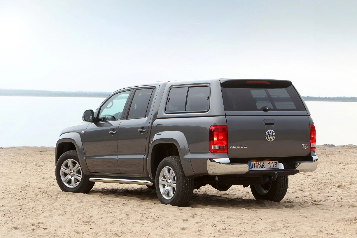 Grau-metallicfarbener VW Amarok mit Hardtop steht am Sandstrand vor einem ruhigen Gewässer unter bewölktem Himmel.