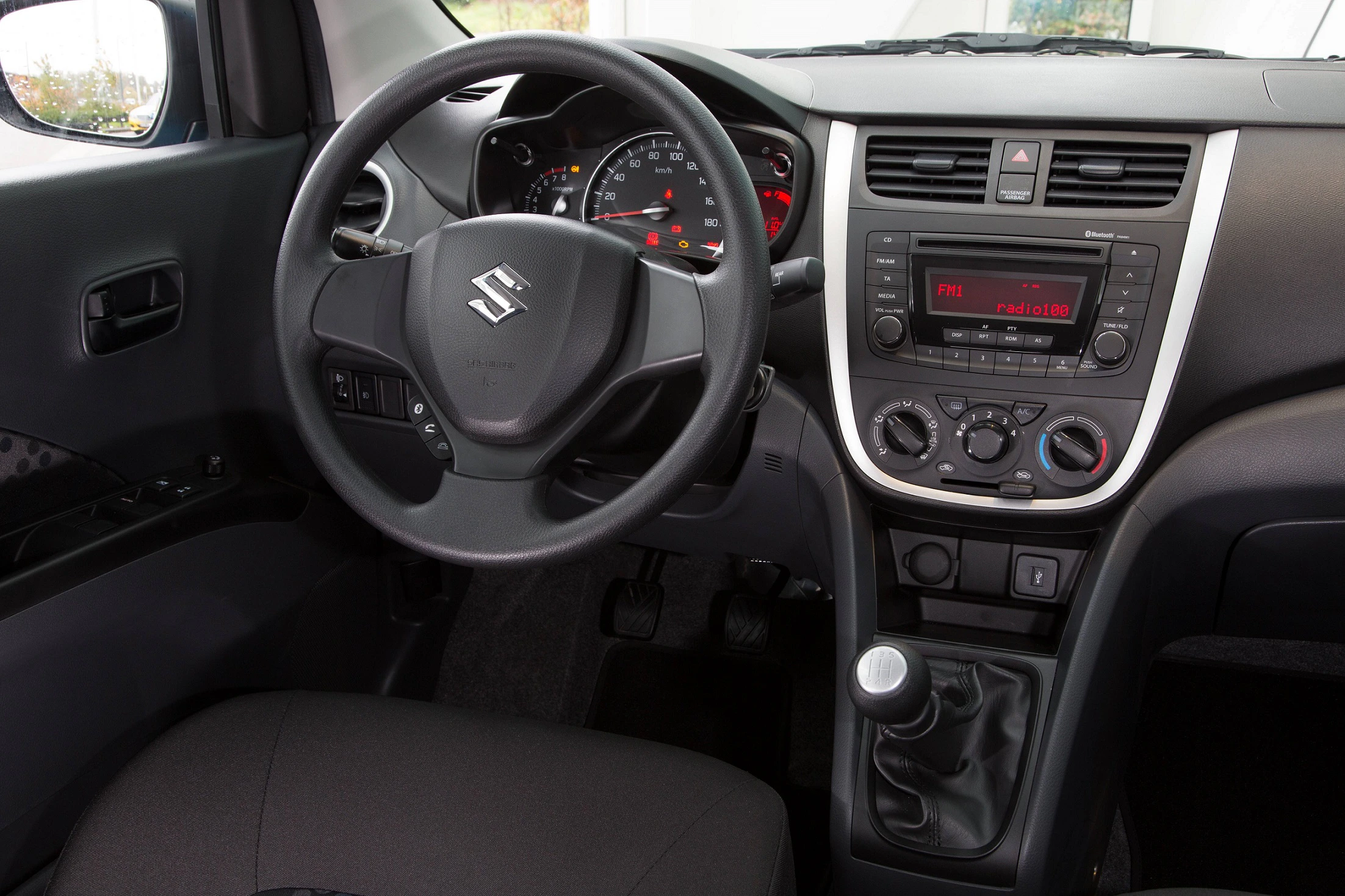 Innenraum eines Suzuki-Autos mit Lenkrad, Tachometer, Klimaanlage und manueller Gangschaltung. Schwarzes Interieur, zentrale Bedienelemente sichtbar.