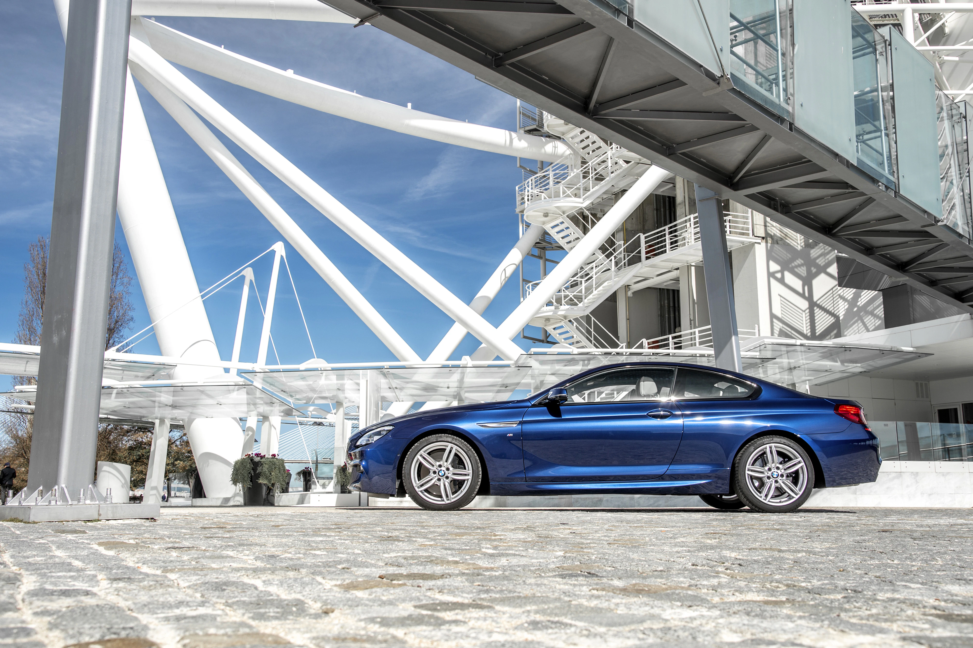 Blaues Auto vor moderner Architektur mit weißen Stahlstrukturen und Glasfassade unter blauem Himmel.