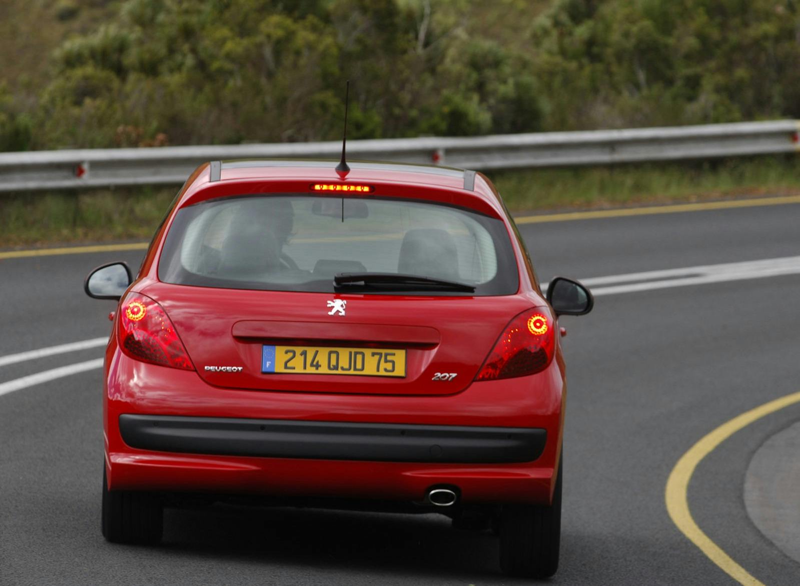 Roter Peugeot 207 auf kurviger Straße, umgeben von grüner Landschaft. Rücklichter leuchten, Fahrzeugkennzeichen sichtbar.